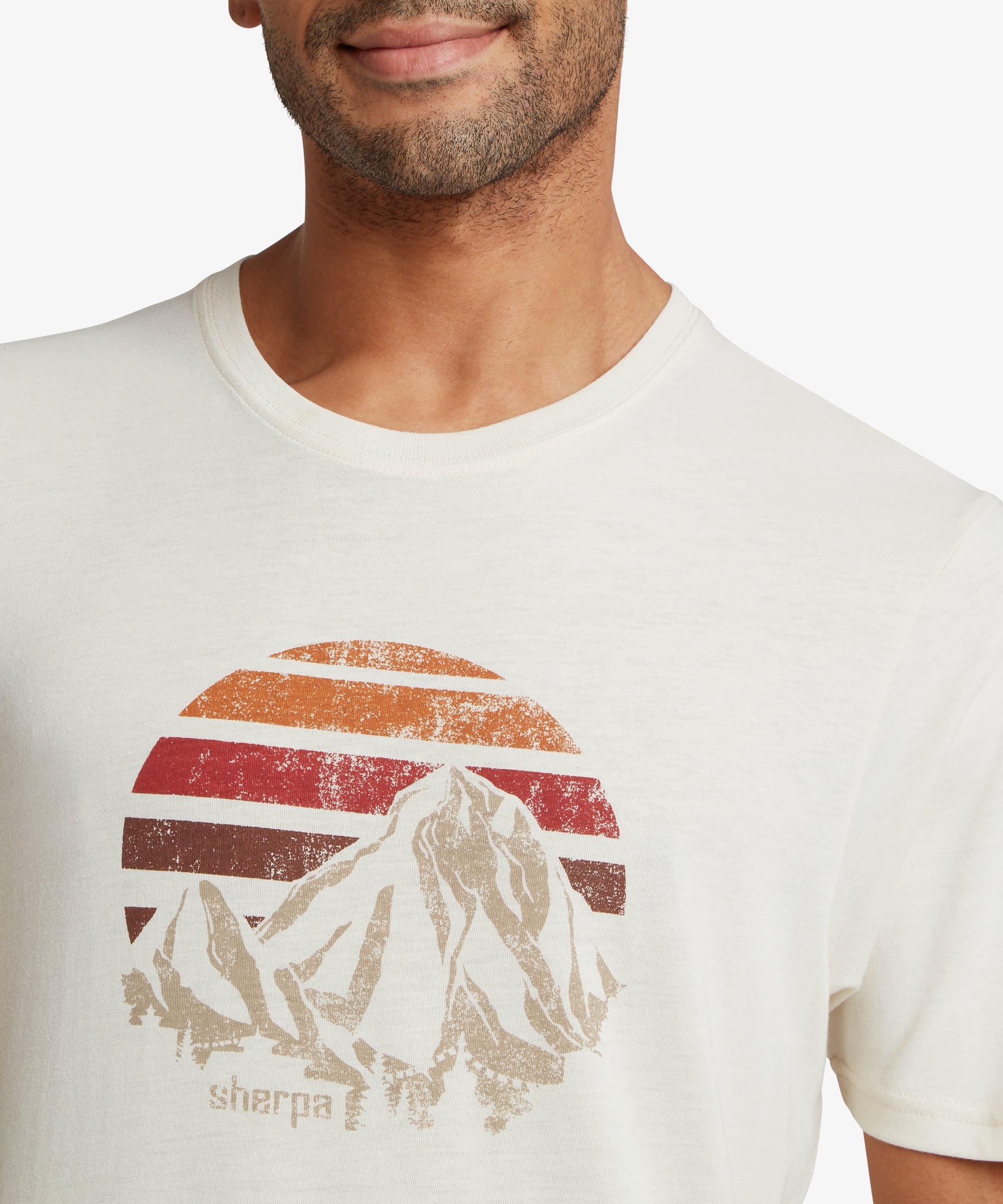 Close‑up of an off‑white T‑shirt showing a circular retro‑style mountain and sunset graphic with red, orange, and brown stripes, along with subtle textured print details.