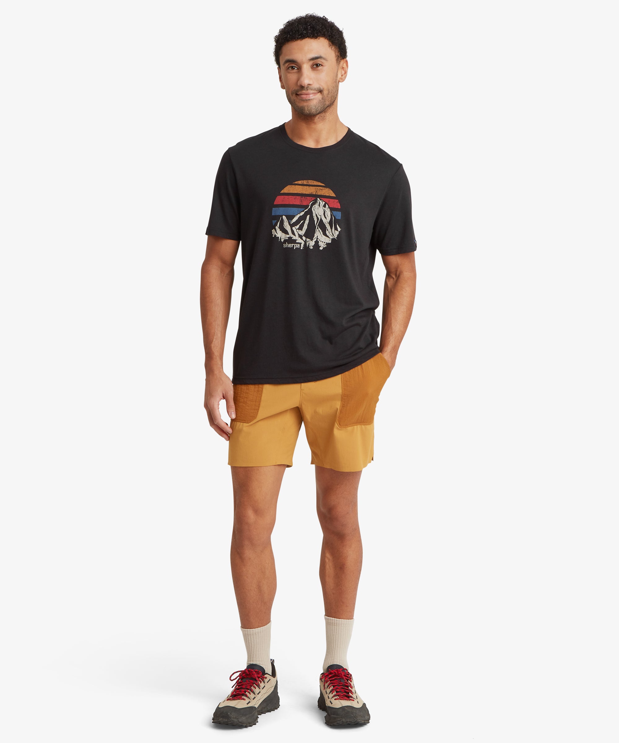 A black T‑shirt with a retro mountain‑and‑sunset chest graphic is worn with burnt orange outdoor shorts, beige socks, and rugged hiking shoes.