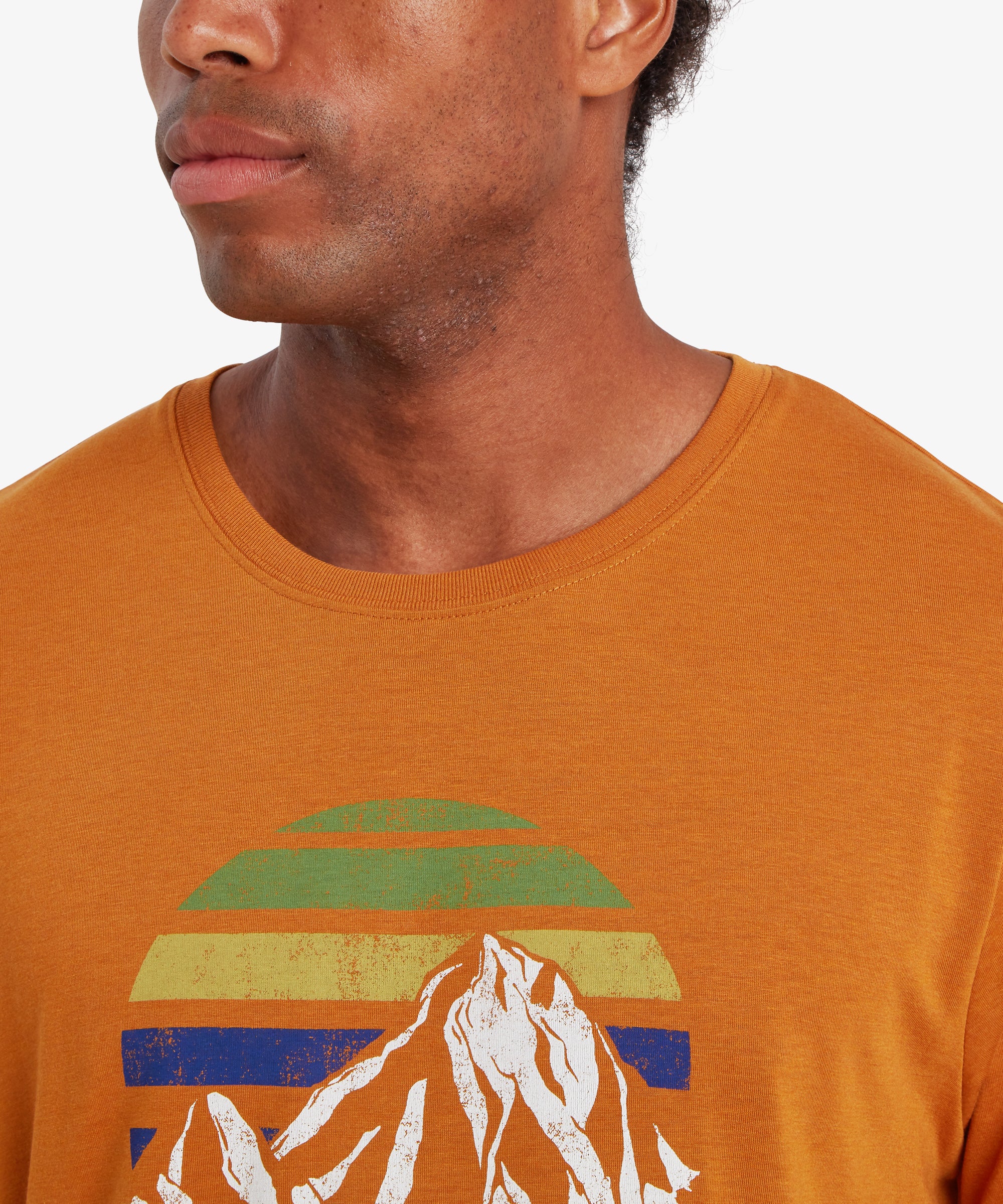 A close up of a man wearing an orange t-shirt with a white mountain and multi-colored sunset graphic design on the chest.