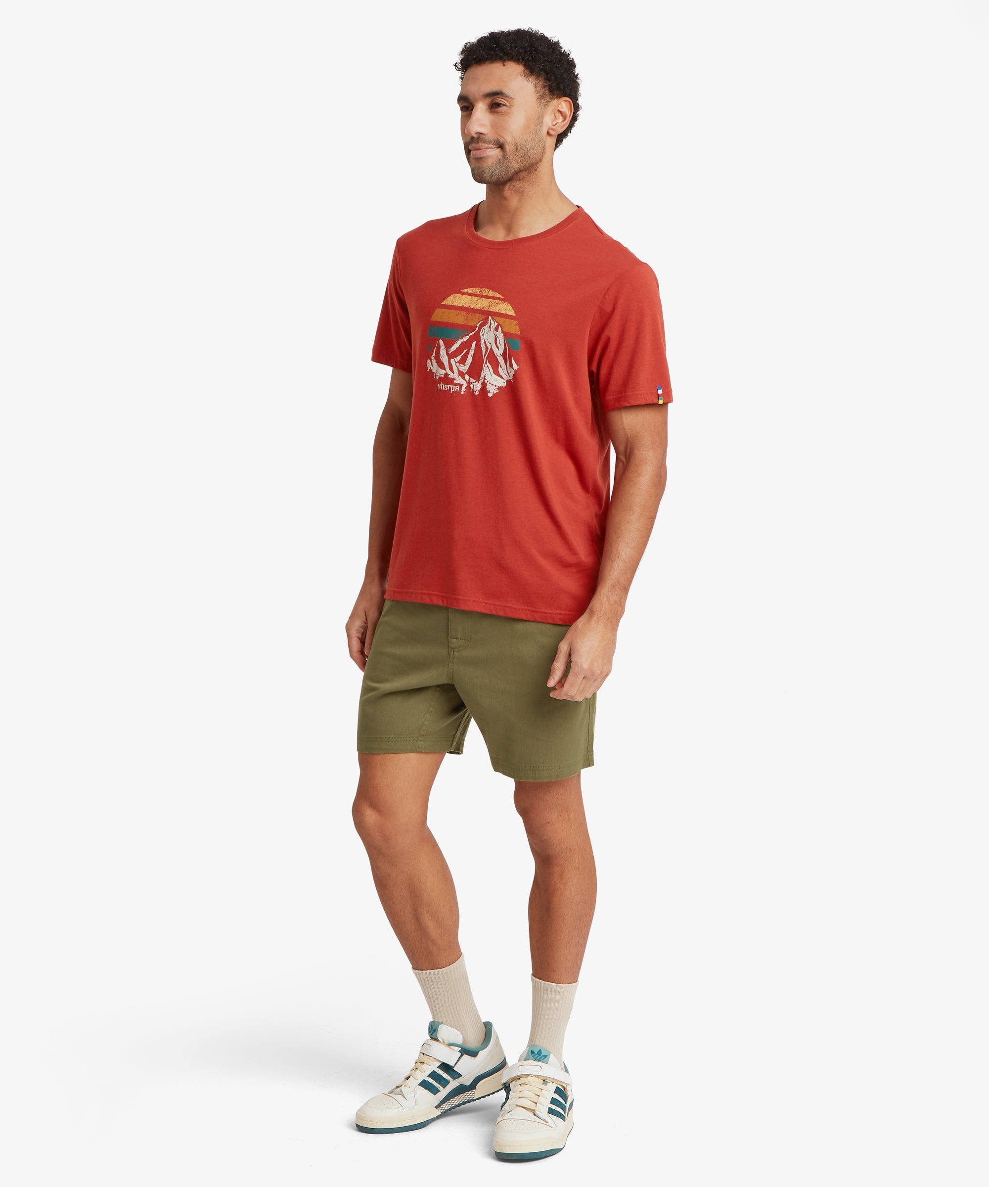 Angled full‑body view of a person wearing a red mountain‑graphic T‑shirt with olive shorts.