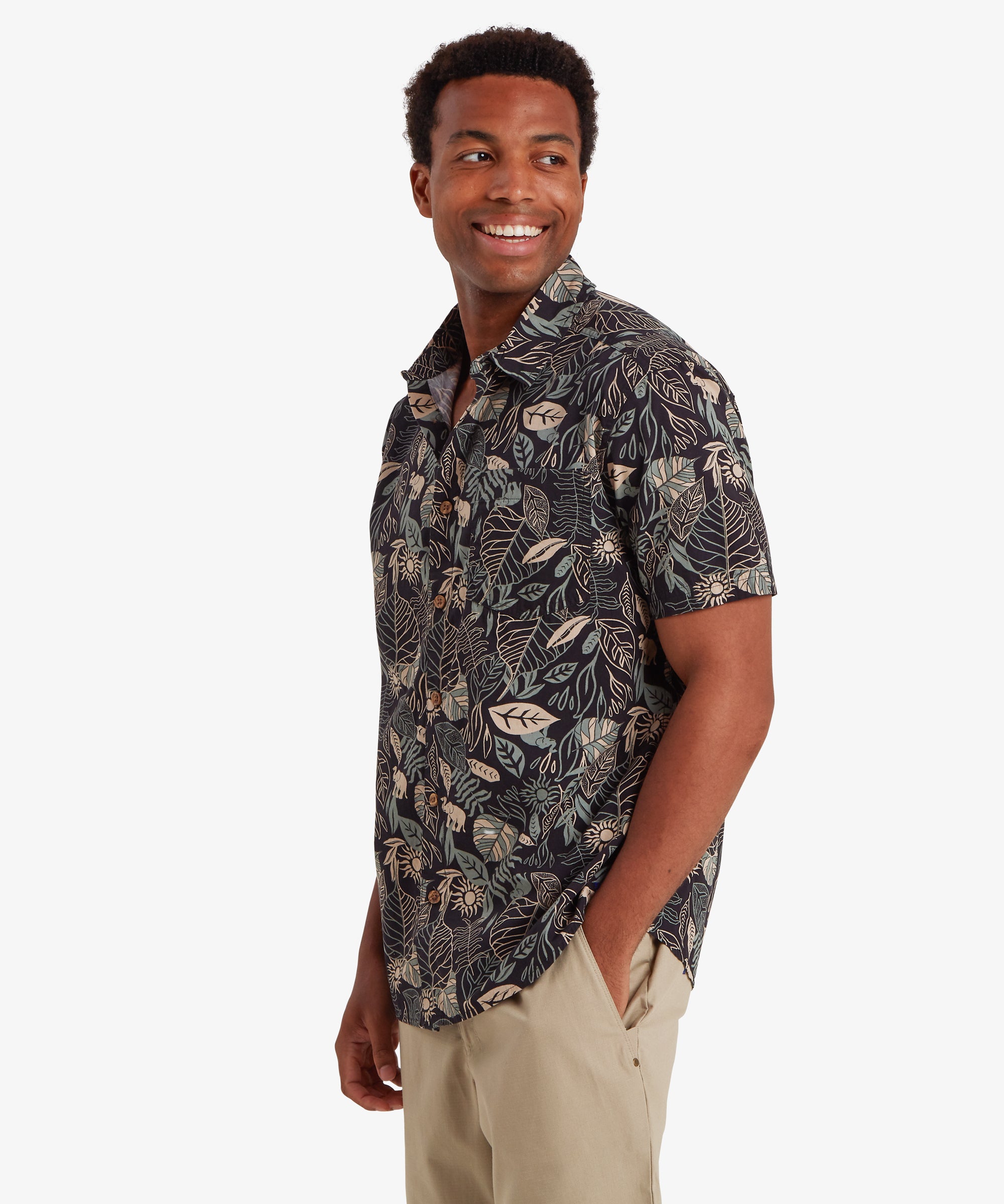 Mann mit schwarzem Kurzarmhemd mit Knšpfen. Das Hemd ist mit einem durchgehenden botanischen Blattmuster in gedeckten Creme- und Blautšnen bedruckt. In Kombination mit beigen Shorts entsteht ein lŠssiger Look fŸr den Alltag. Der Mann hat seinen Kšrper leicht nach rechts gedreht, lŠchelt und schaut leicht hinter sich, wŠhrend seine linke Hand in der Tasche ruht.