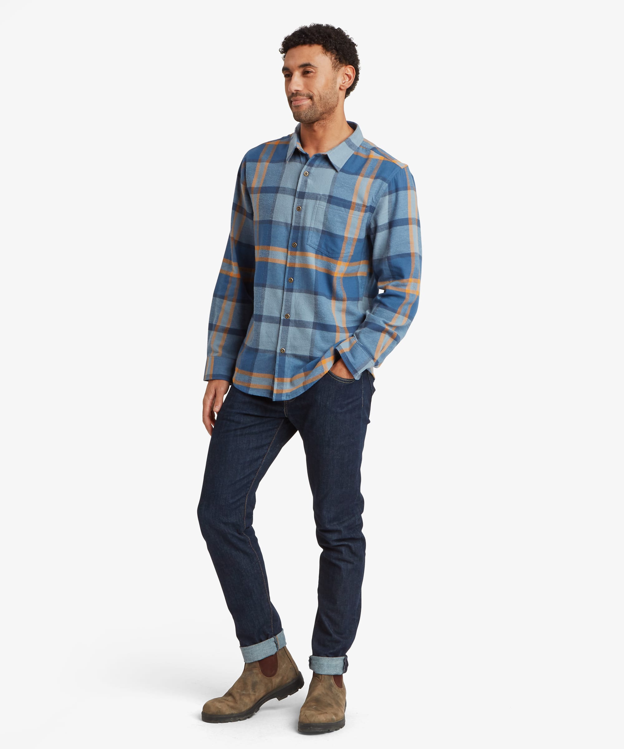 Full-body view of a man standing casually in a blue plaid long-sleeve shirt with a relaxed fit. The shirt features a button-front placket and classic collar and is styled with dark jeans and casual shoes.