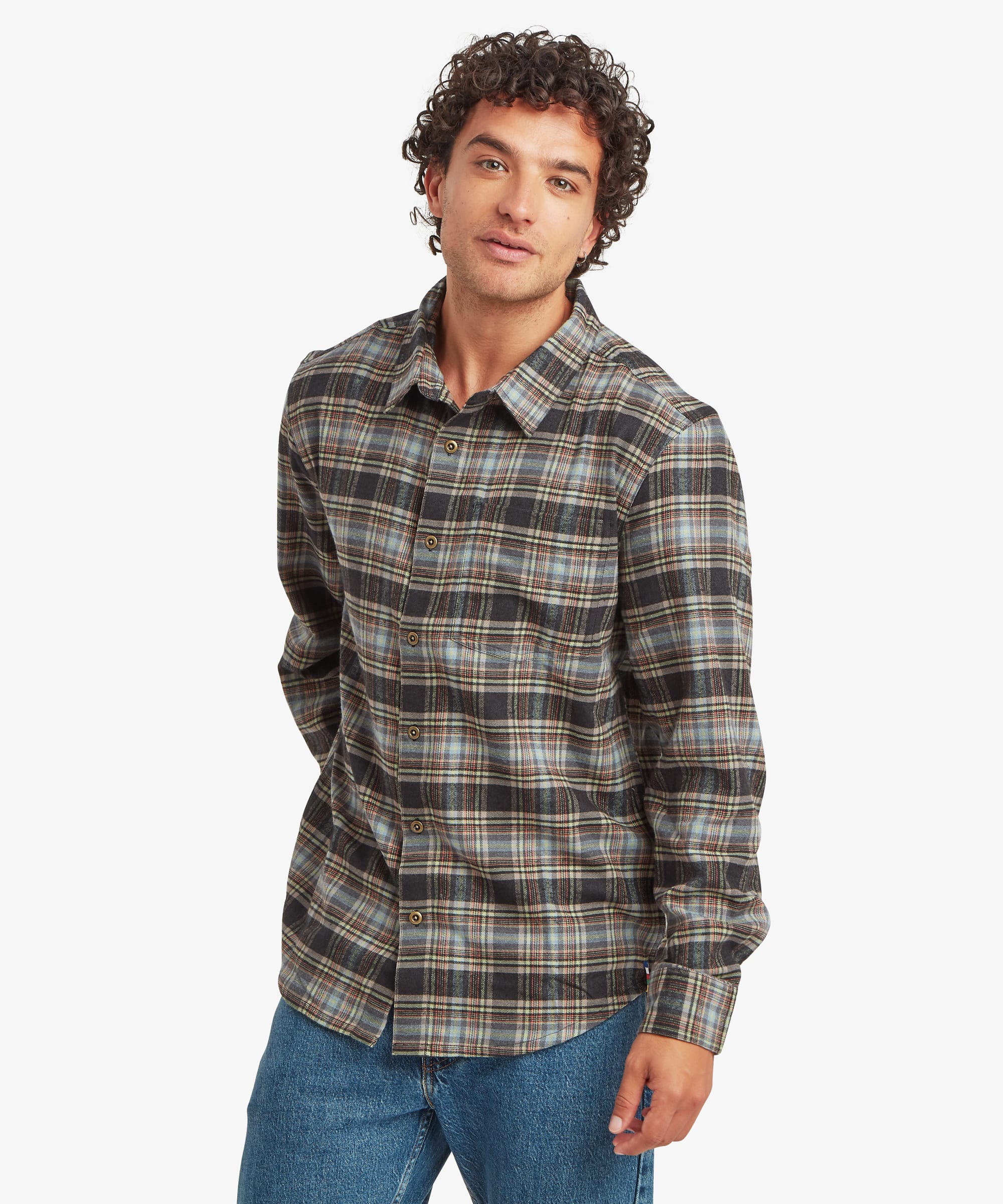 Front view of a man wearing a long-sleeve plaid shirt in olive green, brown, cream and charcoal tones. The shirt has a relaxed regular fit, button-front closure, spread collar and a single chest pocket. The man stands casually with a slight smile, one hand tucked into a pocket.