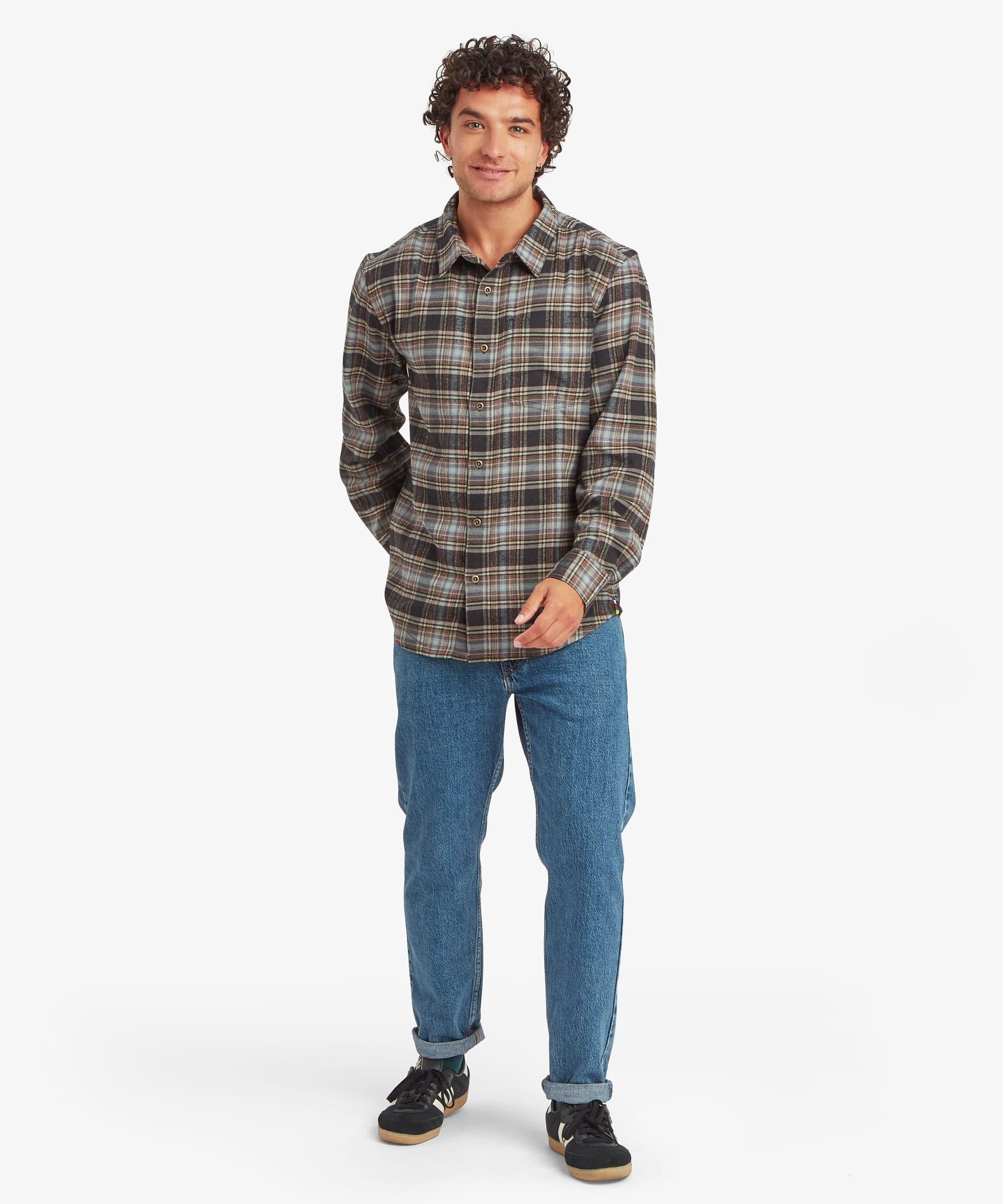 Full-body view of a man wearing an olive, brown and cream plaid long-sleeve shirt paired with blue jeans and casual shoes. The shirt has a relaxed fit through the body, a curved hem, button-front closure, chest pocket and classic collar. One hand rests in a pocket while the other hangs naturally by his side.