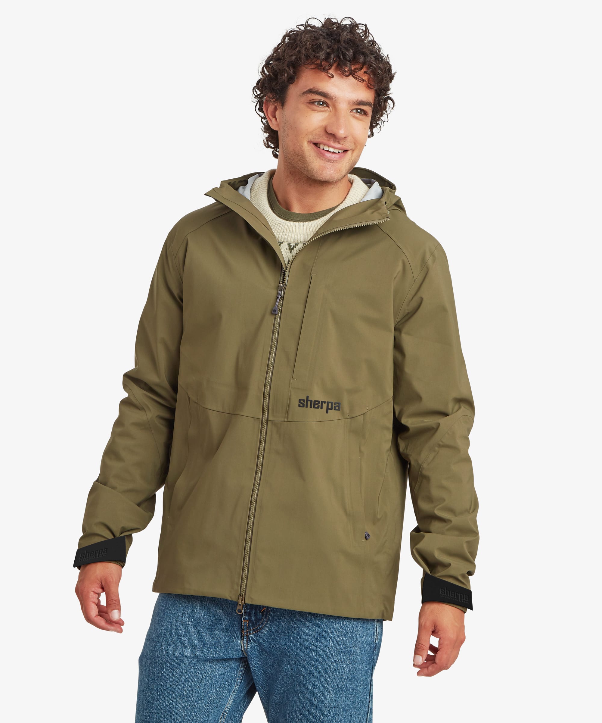A portrait of a man wearing an olive green waterproof, hooded jacket with black cuffs and a black 'Sherpa Adventure Gear' logo on the chest, paired with blue jeans, against a light gray background.