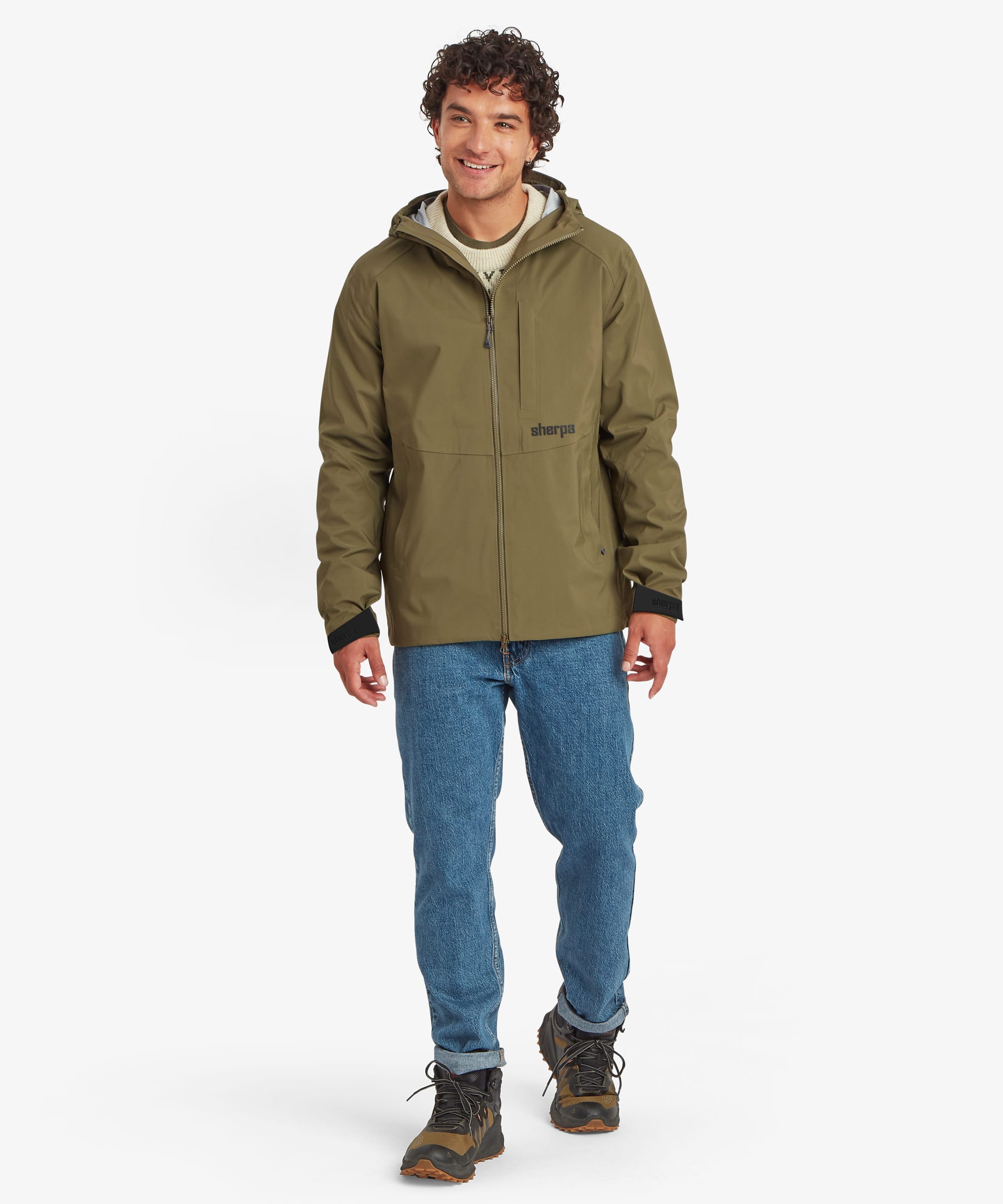 A portrait of a man wearing an olive green Sherpa Adventure Gear, waterproof hooded jacket, blue jeans, and hiking boots, standing against a plain white studio background. The jacket has a small black logo on the left chest that reads "Sherpa"