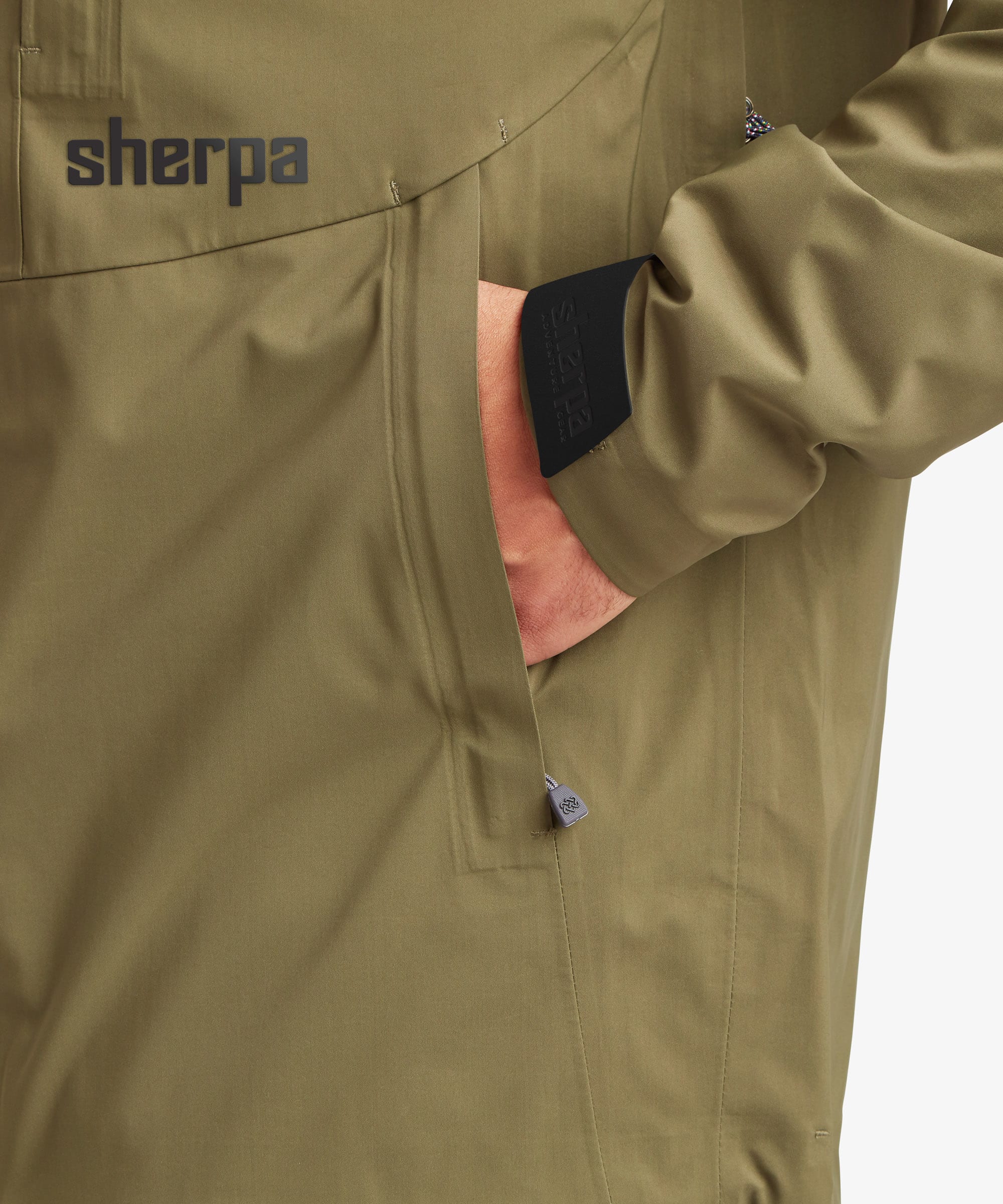 Close-up of a person wearing an olive green Sherpa Adventure Gear waterproof jacket, showing a side pocket with a hand inside and the Sherpa logo on the chest.