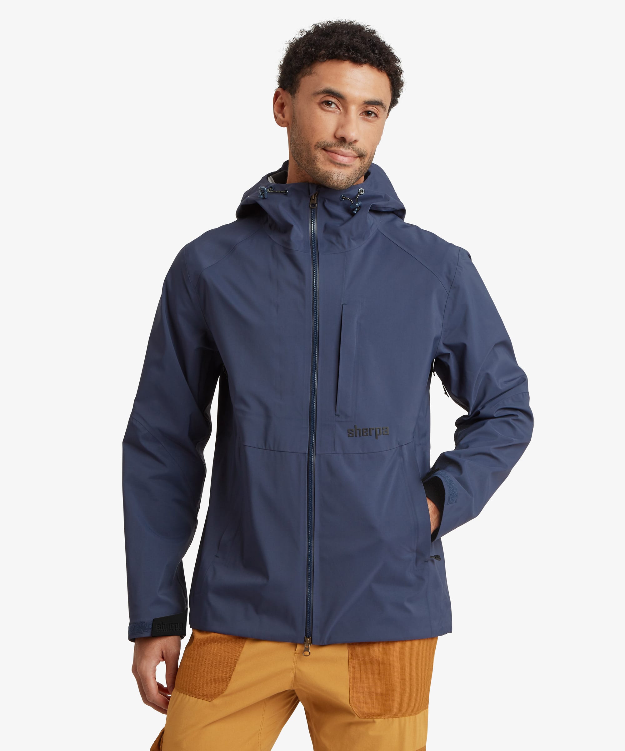 A man wearing a navy blue hooded jacket with a black zipper and a small black 'Sherpa' logo on the waist, paired with mustard yellow pants, against a light gray background.