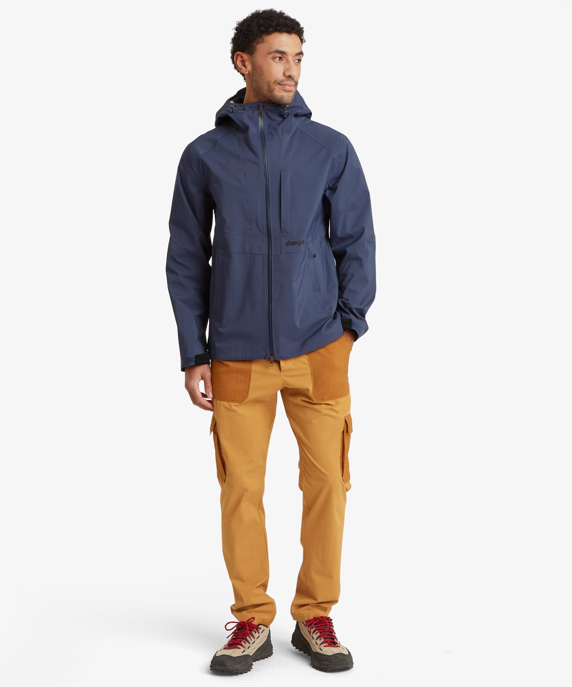 A full length portrait of a man wearing a navy blue hooded waterproof jacket with a black zipper and a small black 'Sherpa' logo on the waist, paired with mustard yellow pants with orange pockets, against a light gray background. The garments are paired with a pair of cream walking shoes.