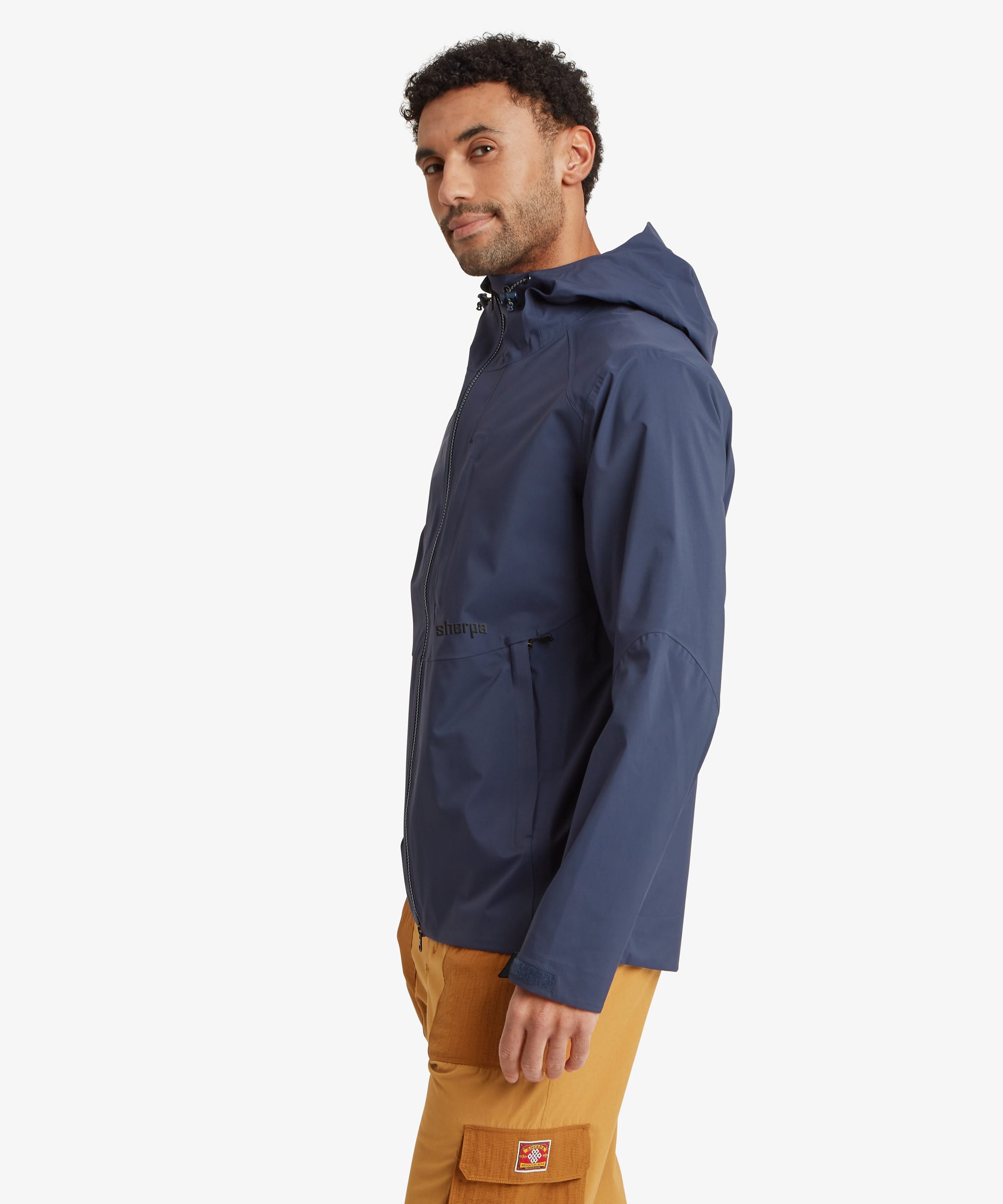 A man wearing a navy blue hooded rain jacket and mustard yellow pants, shown in profile against a light gray studio background. The garments feature the "Sherpa Adventure Gear" logo on the waist and trouser pockets.