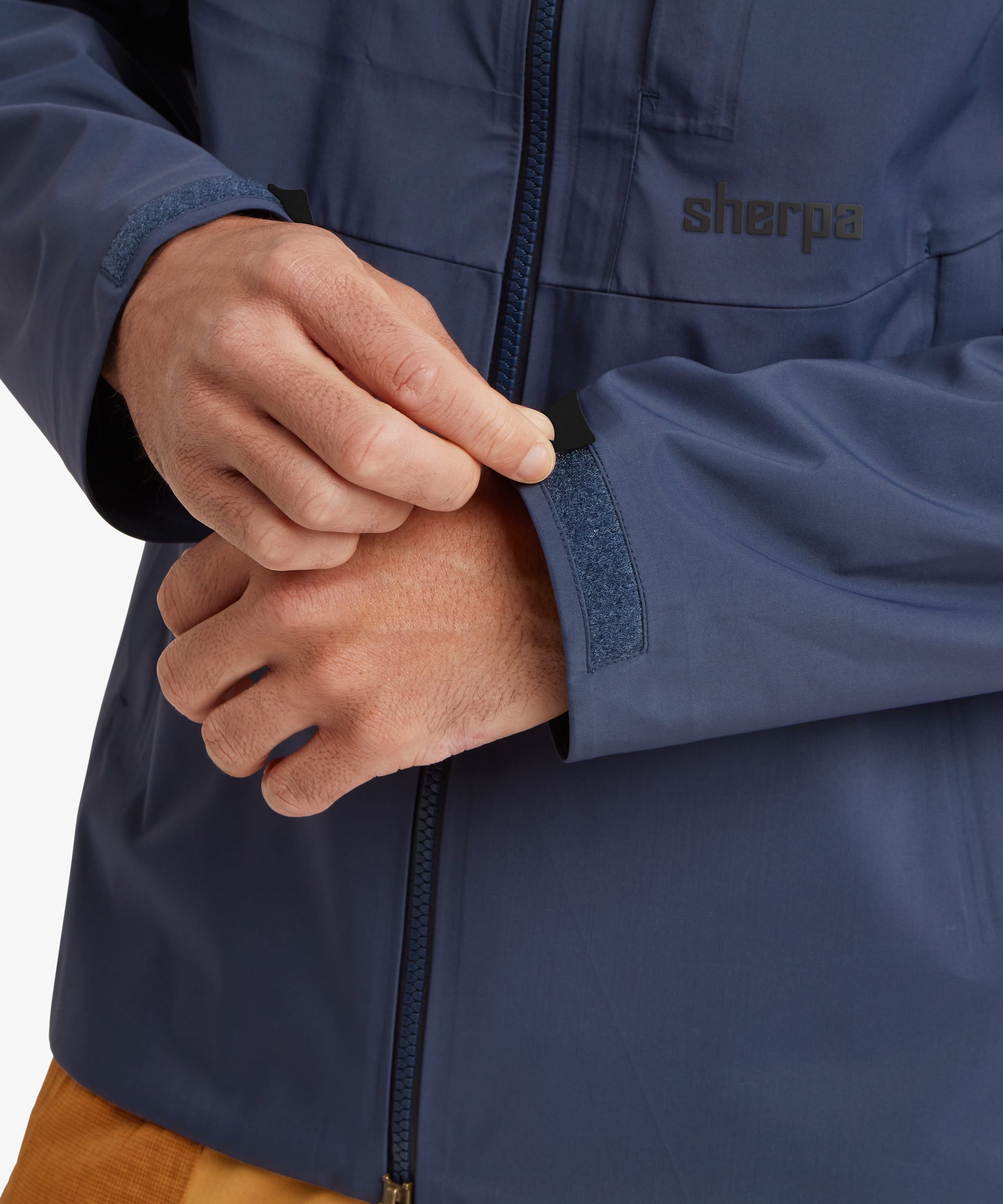 Close-up of a person wearing a navy blue Sherpa Adventure Gear waterproof jacket, showing the brand logo on the chest and the hands adjusting the sleeve cuff.