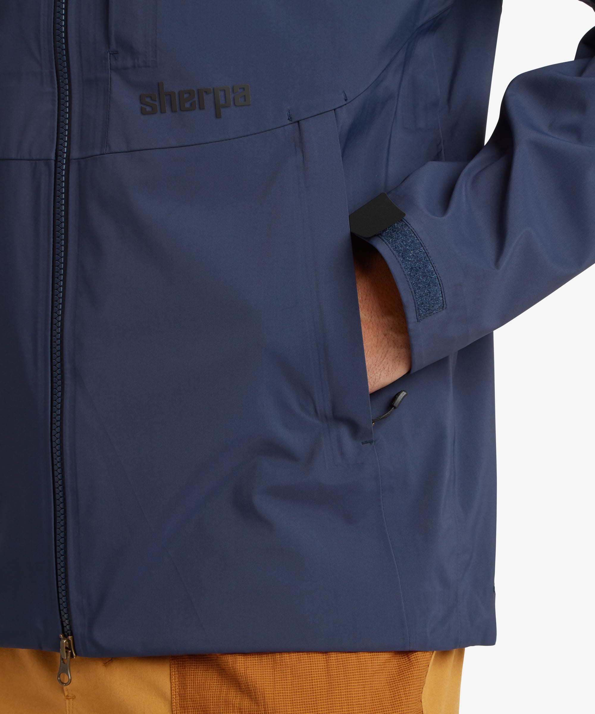 Close-up of a navy blue Sherpa Adventure Gear waterproof Beyul jacket with a front zipper, side pocket showing a hand inside, and the Sherpa logo on the chest. The jacket cuffs feature a Velcro tightening fastening.
