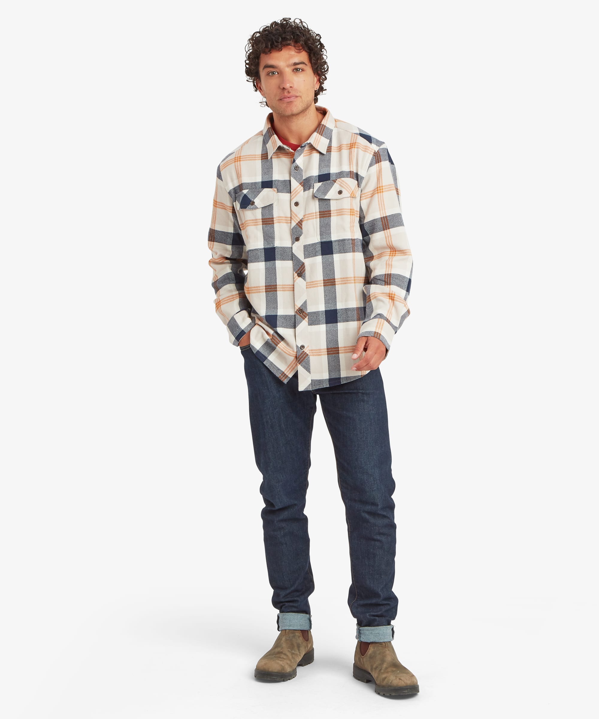 Full-body view of a man wearing a cream plaid overshirt with navy and tan checks, styled with blue jeans and outdoor shoes. The relaxed-fit shirt features two button-flap chest pockets, natural-look buttons and a curved hem. One hand rests in a pocket.