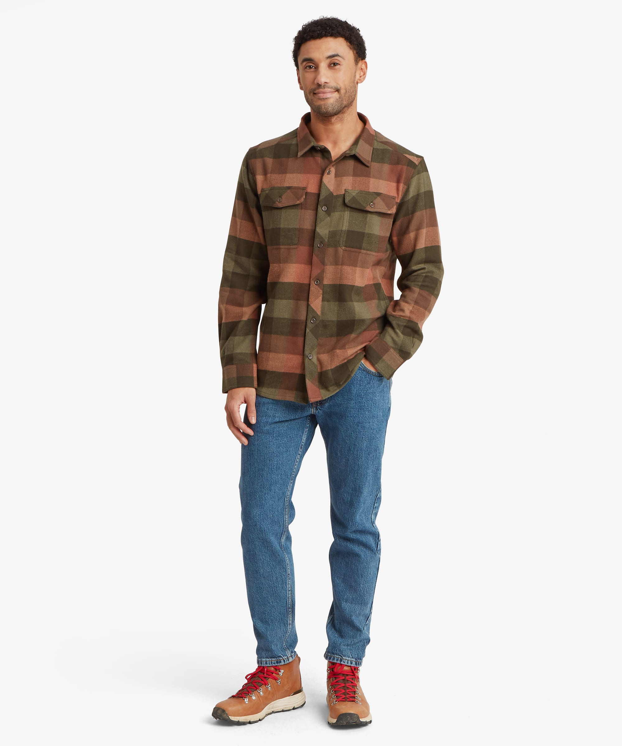 Full-body view of a man wearing an olive green and rust plaid overshirt paired with blue jeans and brown shoes. The shirt features a relaxed fit, button-front closure, curved hem and dual chest pockets with button flaps.