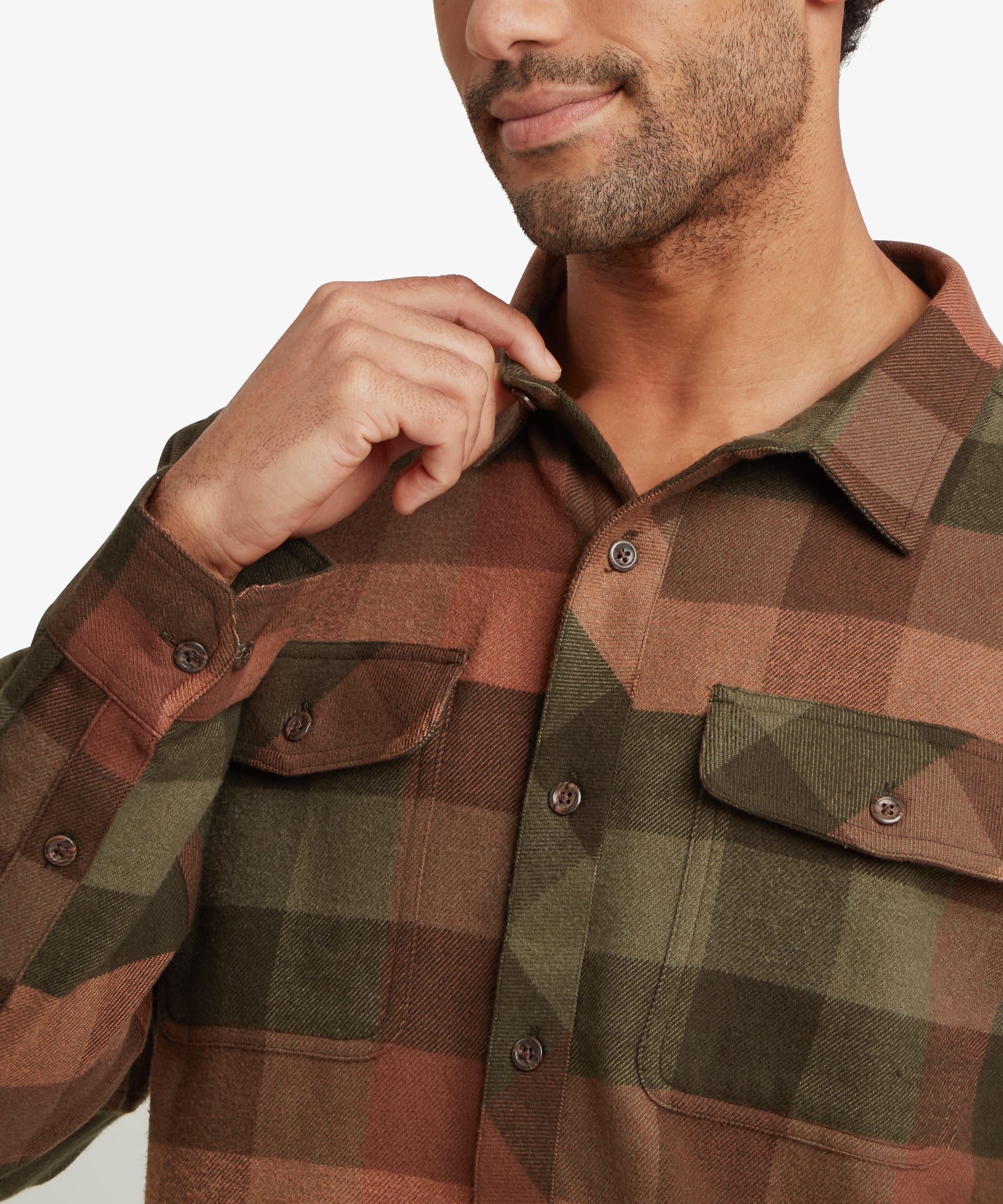 Close-up of the upper chest and collar of a men's plaid overshirt in olive green, rust brown and charcoal tones. Features include a spread collar, natural-look button closure and two button-flap chest pockets. The large-scale plaid pattern and soft brushed fabric texture are clearly visible.