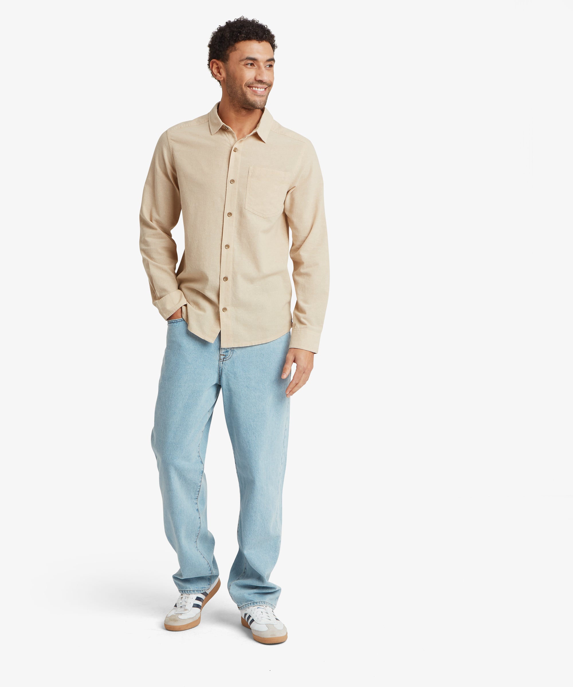 A full-length view of a long-sleeve beige button‑up shirt with a chest pocket, worn untucked with light blue straight-leg jeans and white trainers with gum soles. One hand is in the pocket, highlighting the relaxed fit.