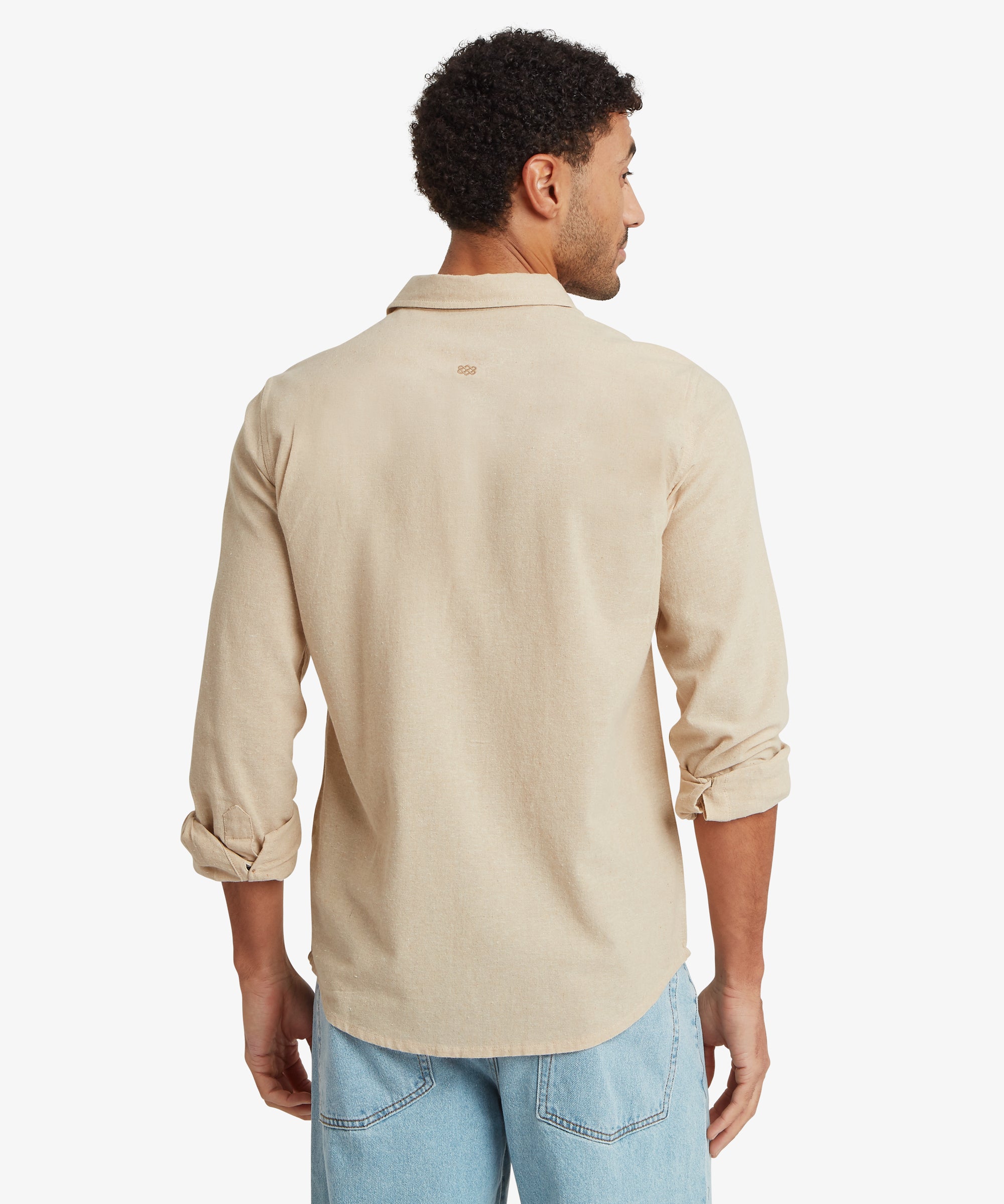 A back view of the beige long‑sleeve button‑up shirt, showing the smooth back panel, curved hemline, and sleeves rolled to mid‑forearm. Light blue jeans complete the outfit.