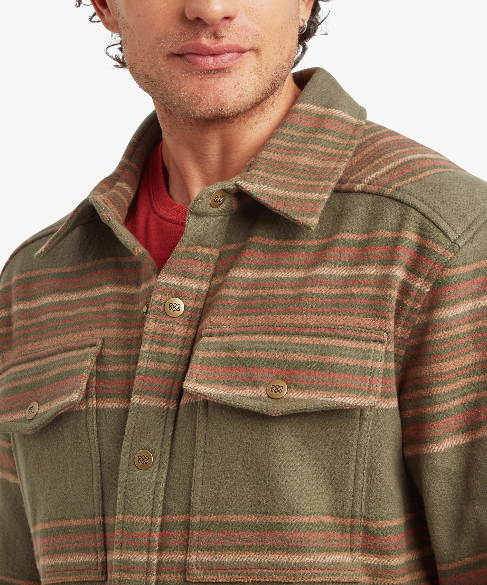 Close-up of the shirt jacket collar and chest area showing natural-look buttons, a spread collar and dual chest pockets with button flaps. The woven stripe pattern combines olive green, rust, tan and charcoal tones. A red T-shirt is visible underneath.