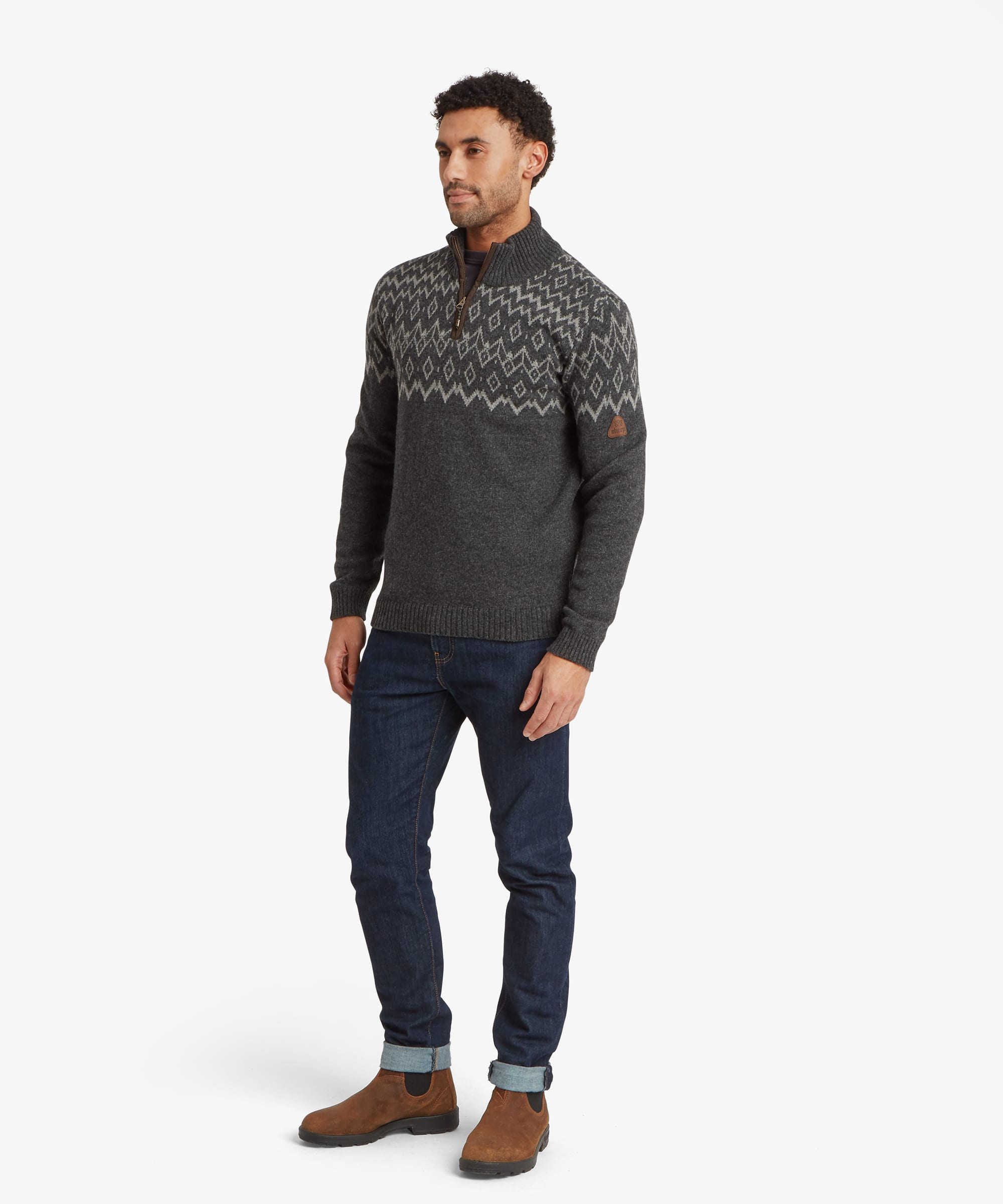 Full-body view of a man wearing a charcoal grey quarter-zip knit sweater paired with blue jeans and brown shoes. The sweater has a regular fit, ribbed cuffs and hem, a high collar and an intricate geometric knit pattern across the upper body.