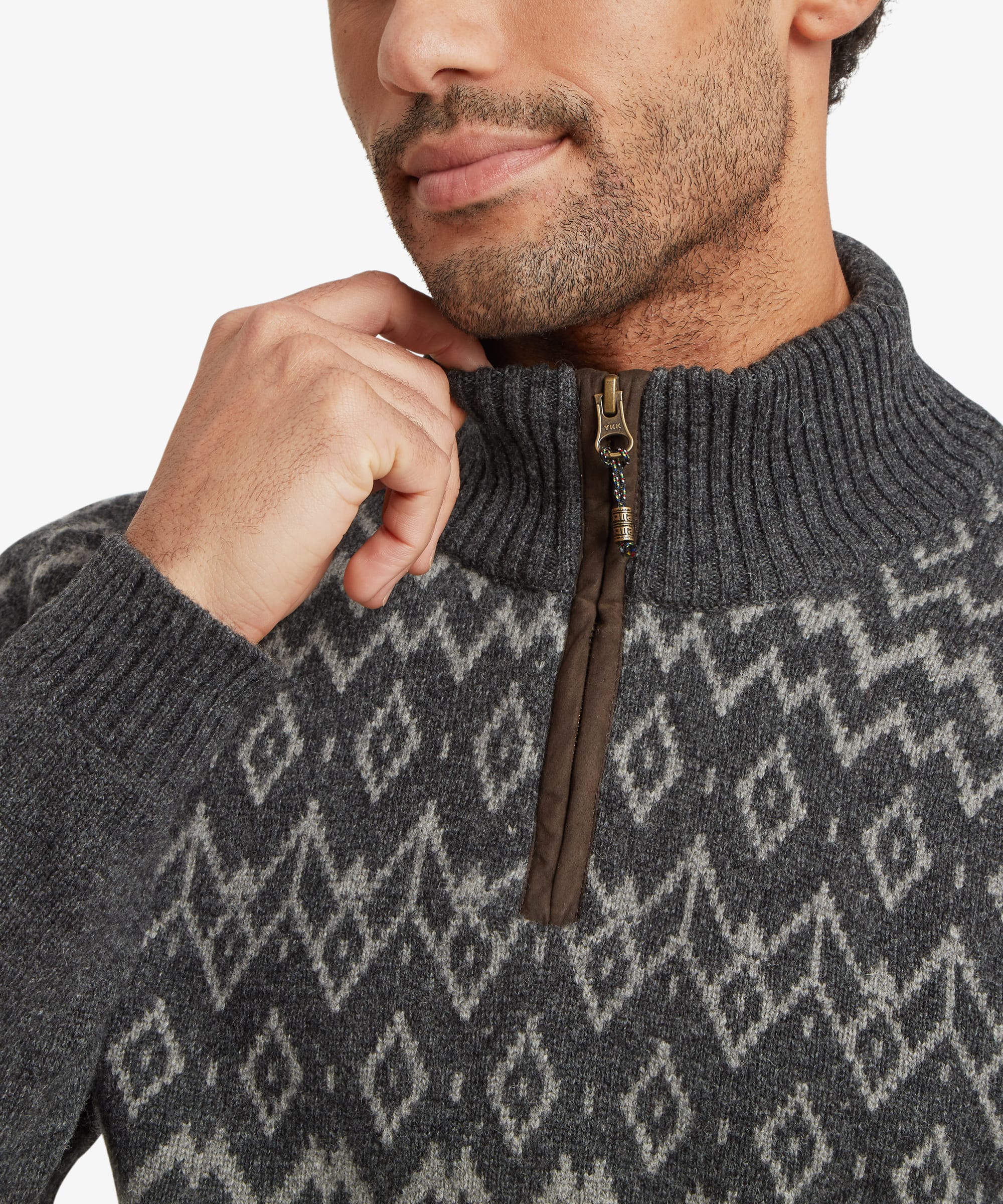 Close-up of the high stand collar and quarter-zip opening on a charcoal grey knit sweater. The geometric knit pattern in grey and cream surrounds the neckline, while the contrast zip tape and puller add detail. The man lightly holds the collar.