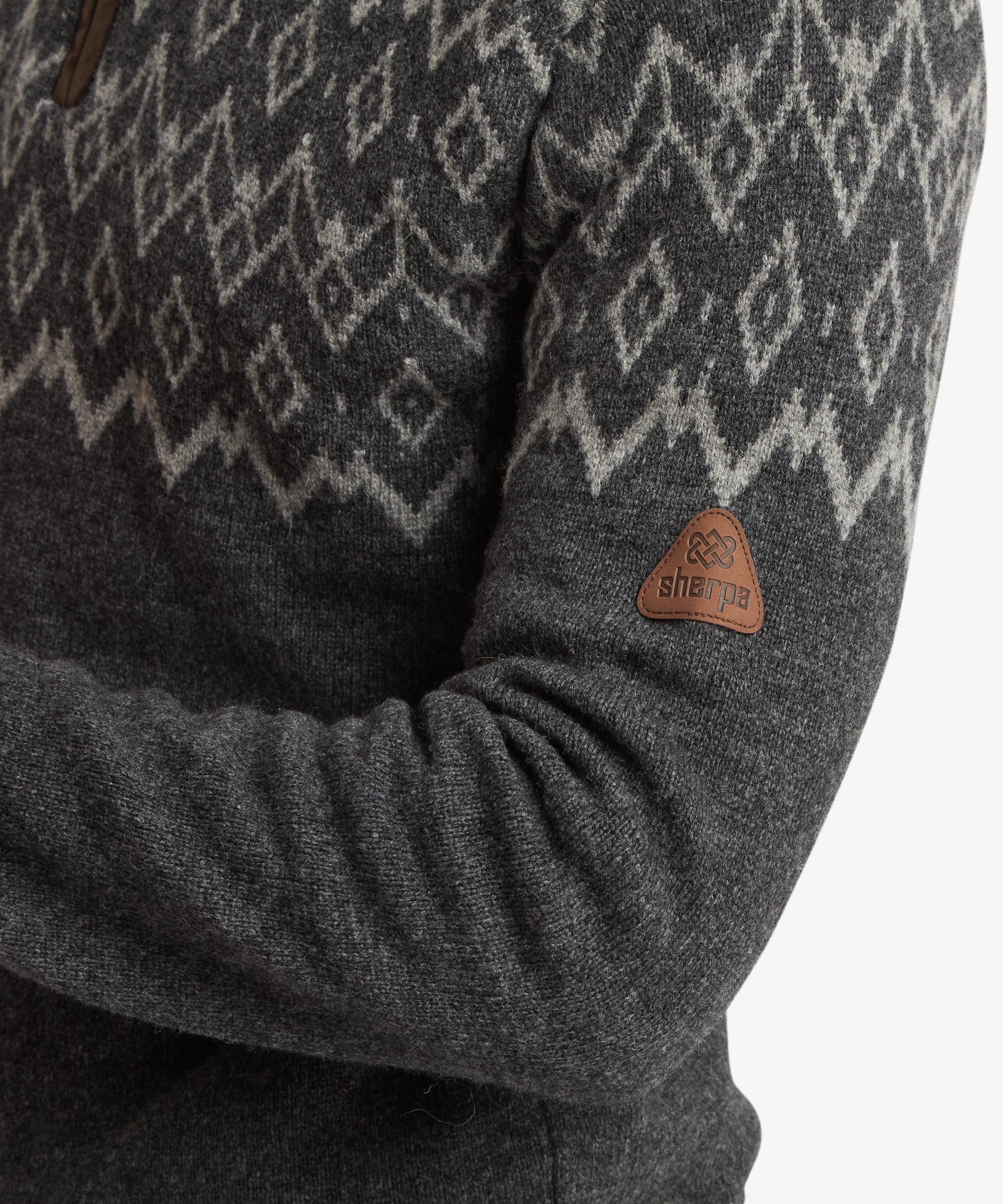 Close-up of the sleeve and shoulder area of the charcoal grey quarter-zip sweater. The image highlights the textured knit construction, geometric Fair Isle-inspired pattern and a small Sherpa logo patch positioned on the upper sleeve.