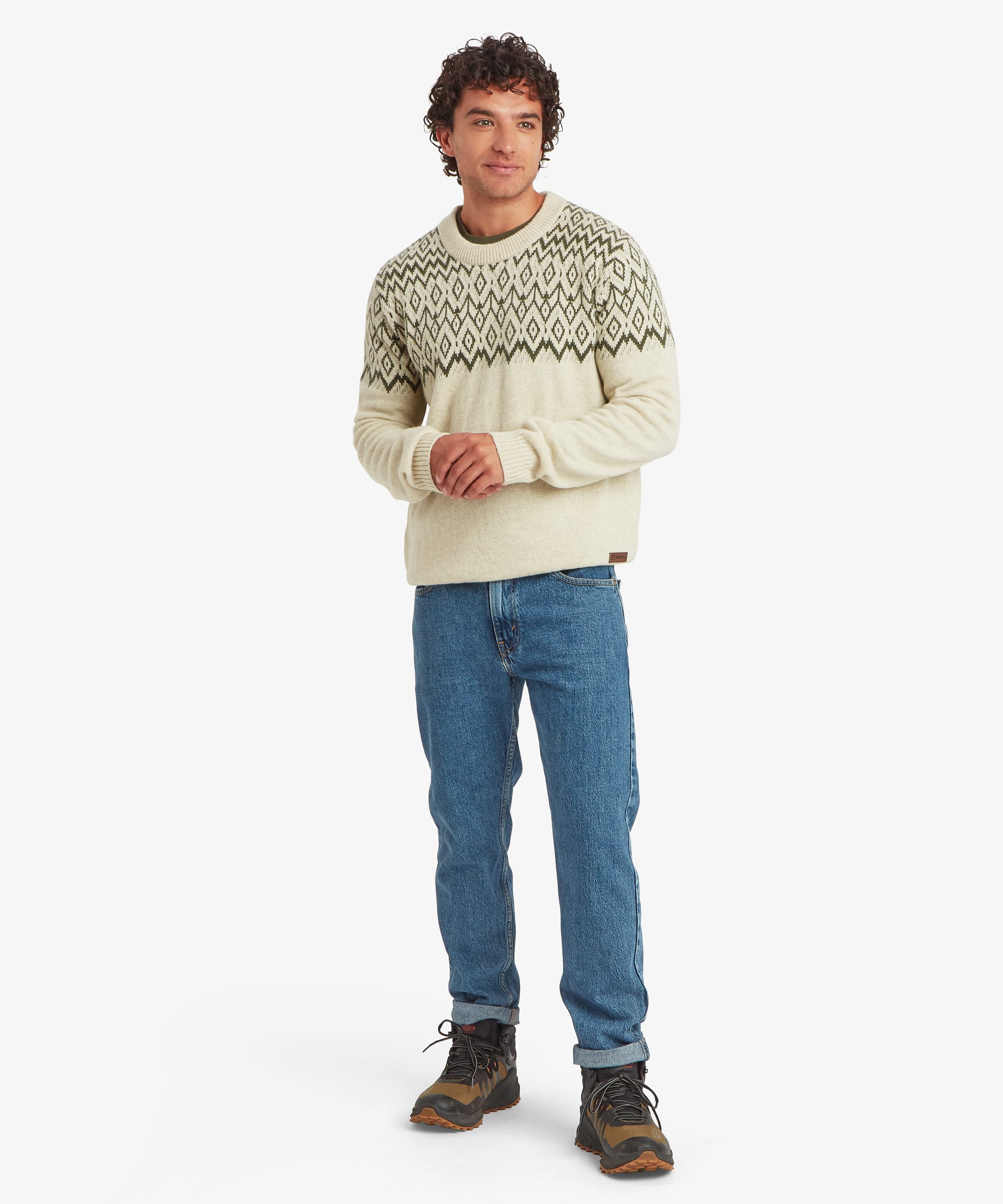 Full-body view of a man wearing a cream crew-neck sweater with olive green geometric knit detailing across the upper body. The regular-fit sweater is paired with blue jeans and outdoor shoes and features ribbed cuffs, hem and neckline.