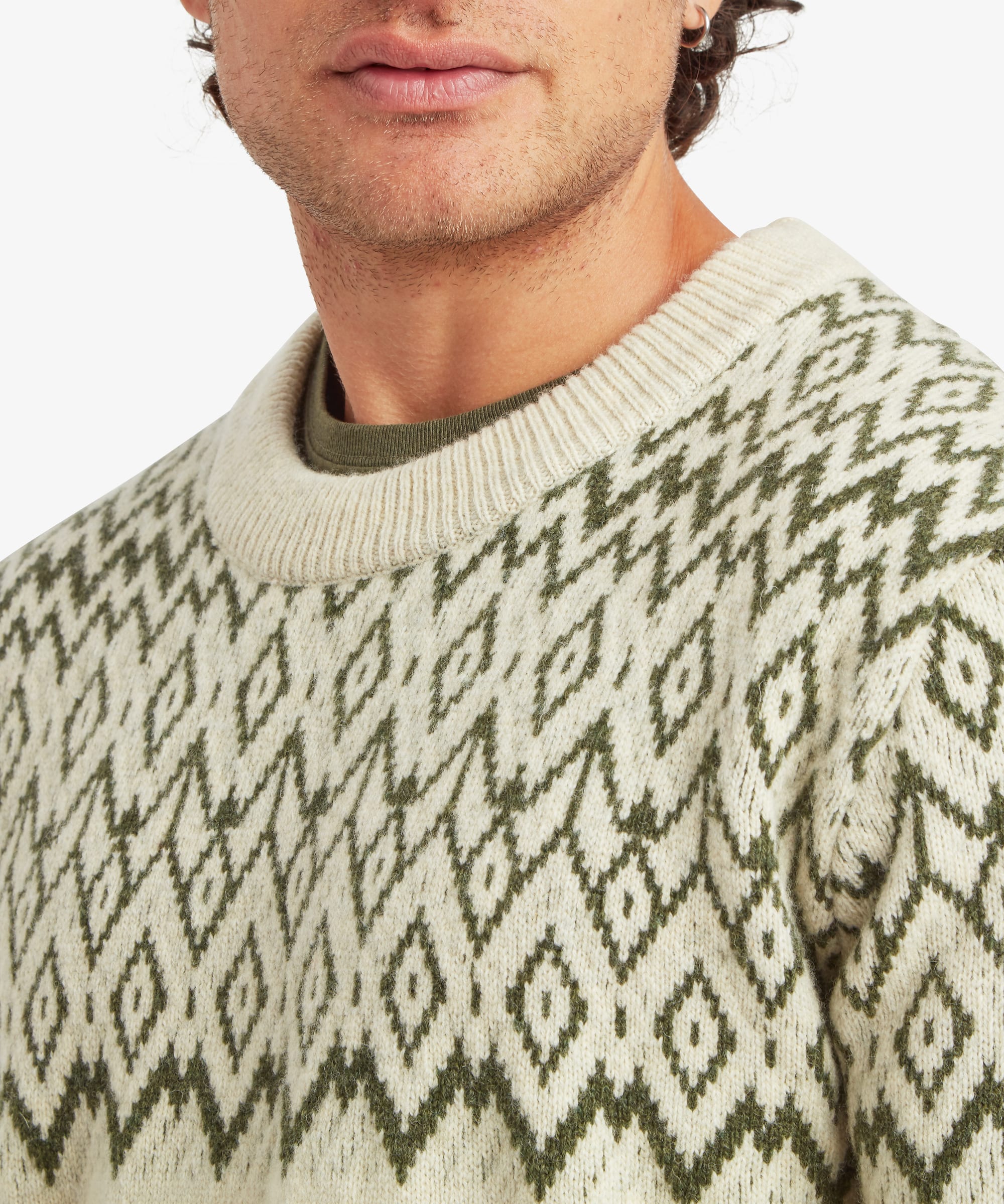 Close-up of the upper chest and shoulder area of a cream crew neck sweater featuring an intricate olive and charcoal geometric diamond pattern knitted across the yoke. Ribbed neckline detail and textured knit construction are shown clearly.