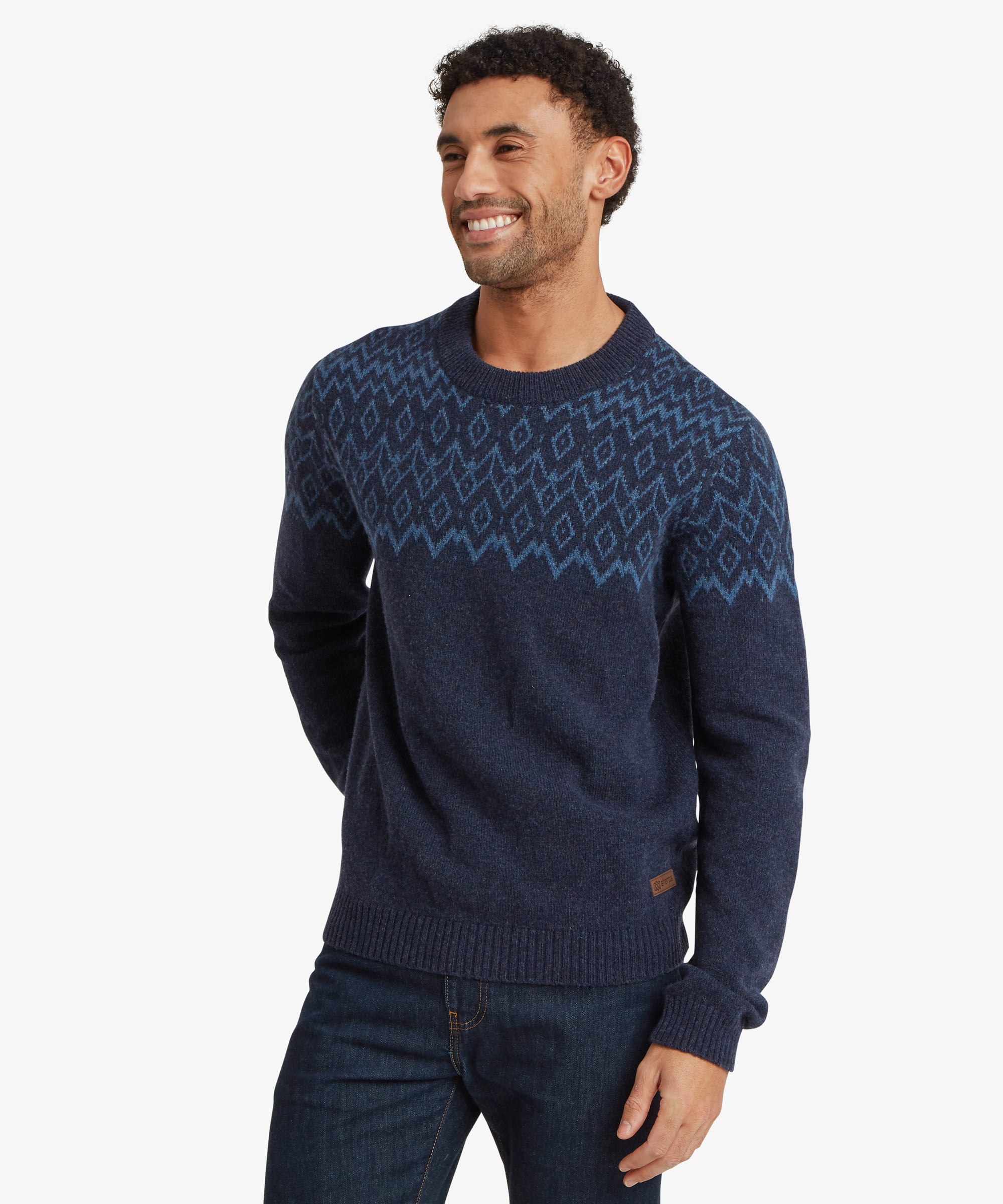 Man smiling while wearing a navy blue crew neck sweater with a tonal blue geometric Fair Isle-inspired pattern across the shoulders and upper chest. Styled with blue jeans, the regular-fit sweater features ribbed cuffs, hem and neckline and offers a comfortable everyday fit.
