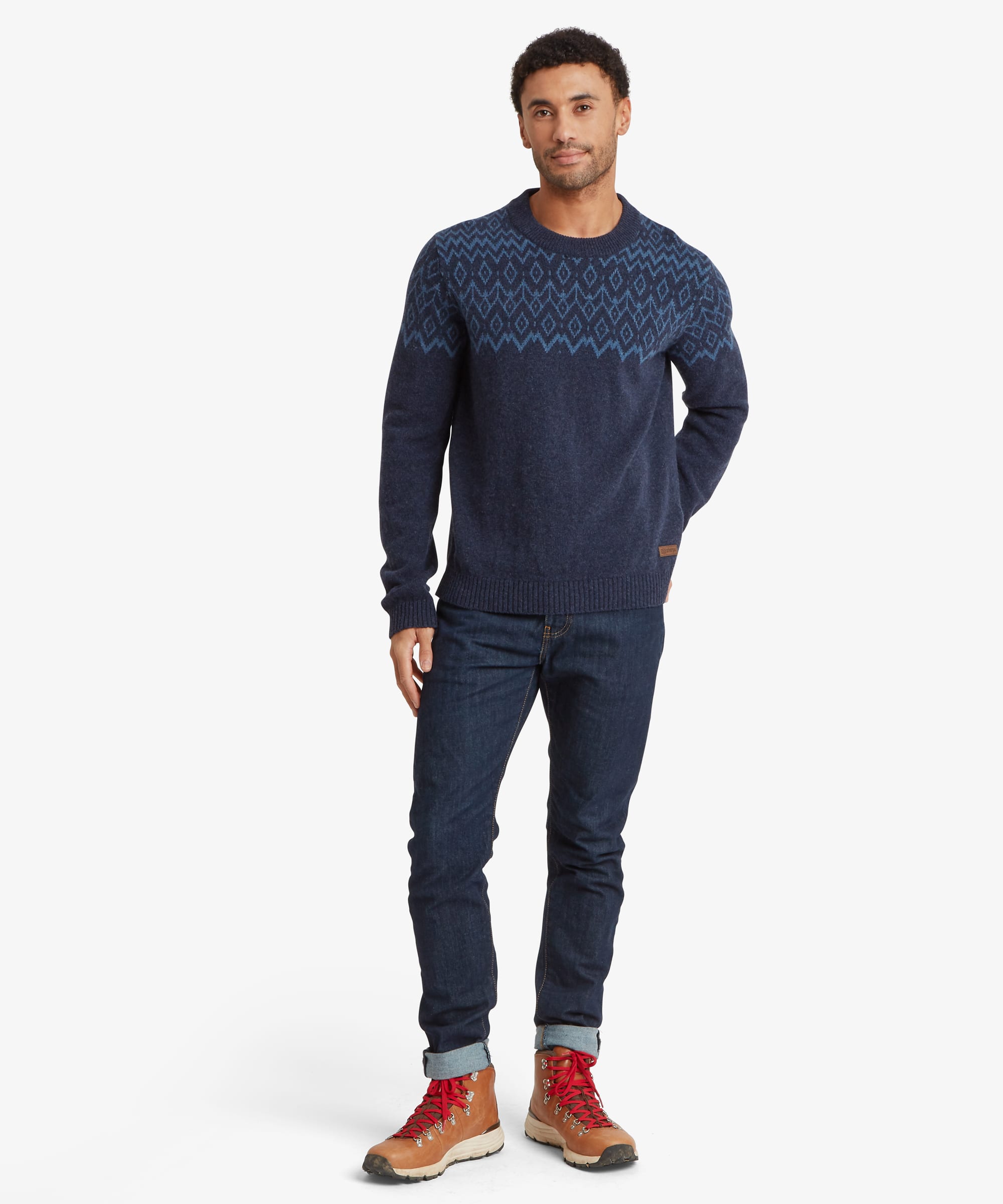 Full-length front view of a man wearing a navy blue crew neck sweater with a blue geometric knit pattern across the upper chest and sleeves. Paired with blue jeans and brown shoes, the sweater has a regular fit with ribbed finishing at the neck, cuffs and hem.