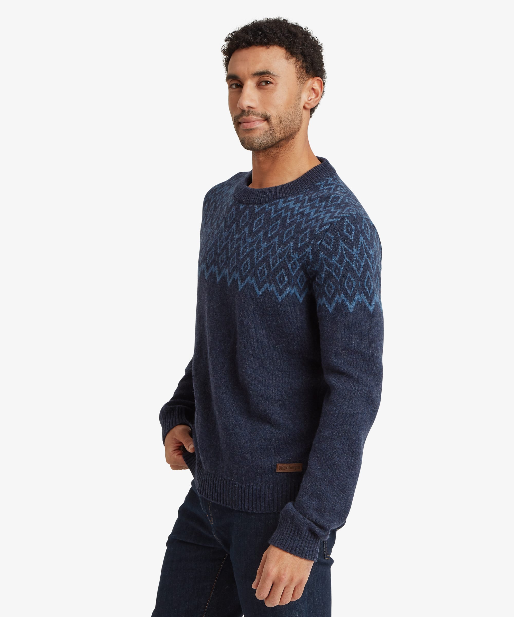 Side profile of a man wearing a navy crew neck sweater with a tonal blue geometric patterned yoke. The regular-fit knit falls straight through the body and sleeves and is paired with blue jeans. The model stands with one arm relaxed by his side.