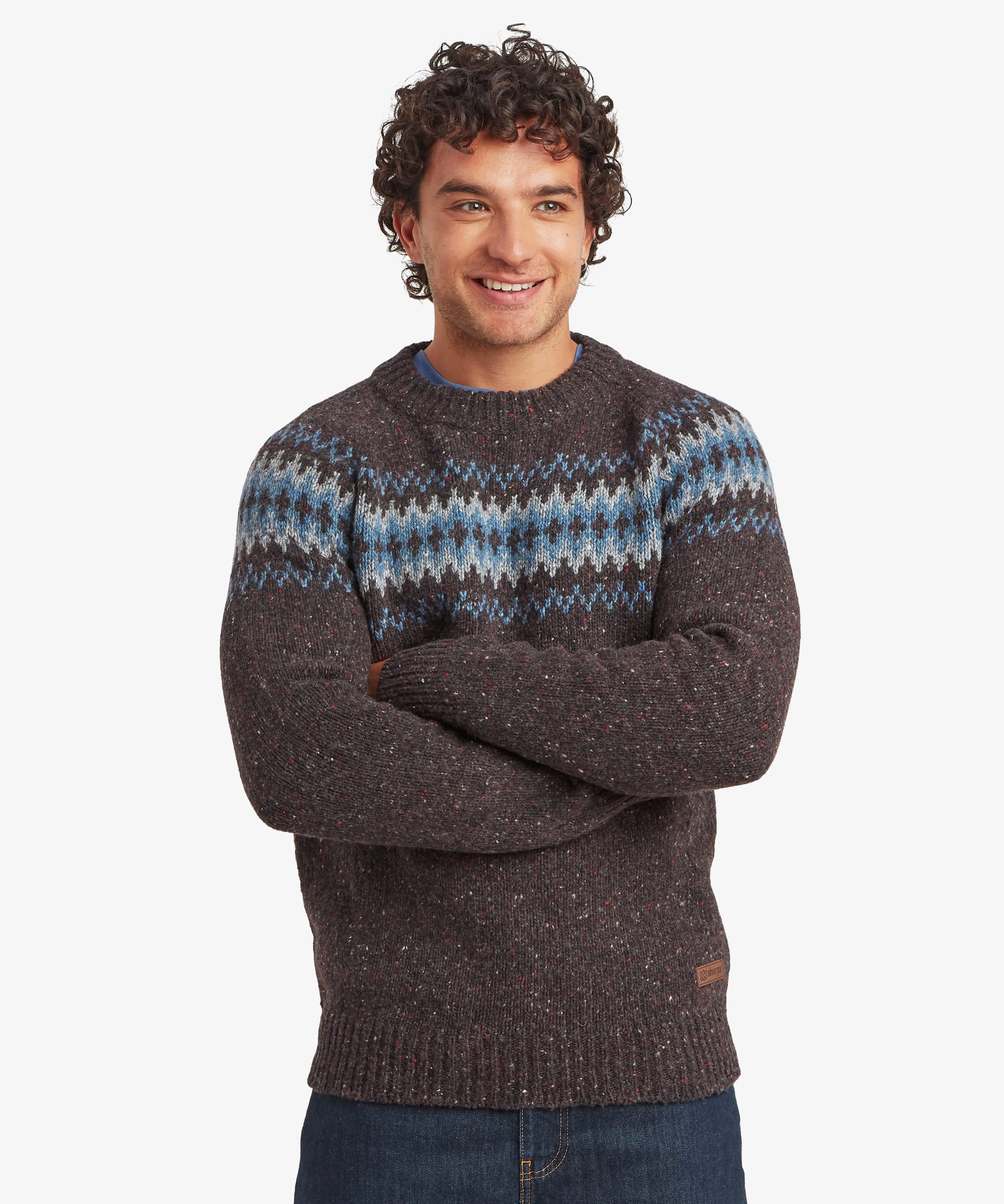 Man smiling with arms folded while wearing a charcoal black crew neck knit sweater featuring a blue, grey and white Fair Isle-inspired geometric pattern across the chest, shoulders and upper sleeves. The regular-fit sweater is paired with blue jeans and finished with ribbed cuffs, hem and neckline.