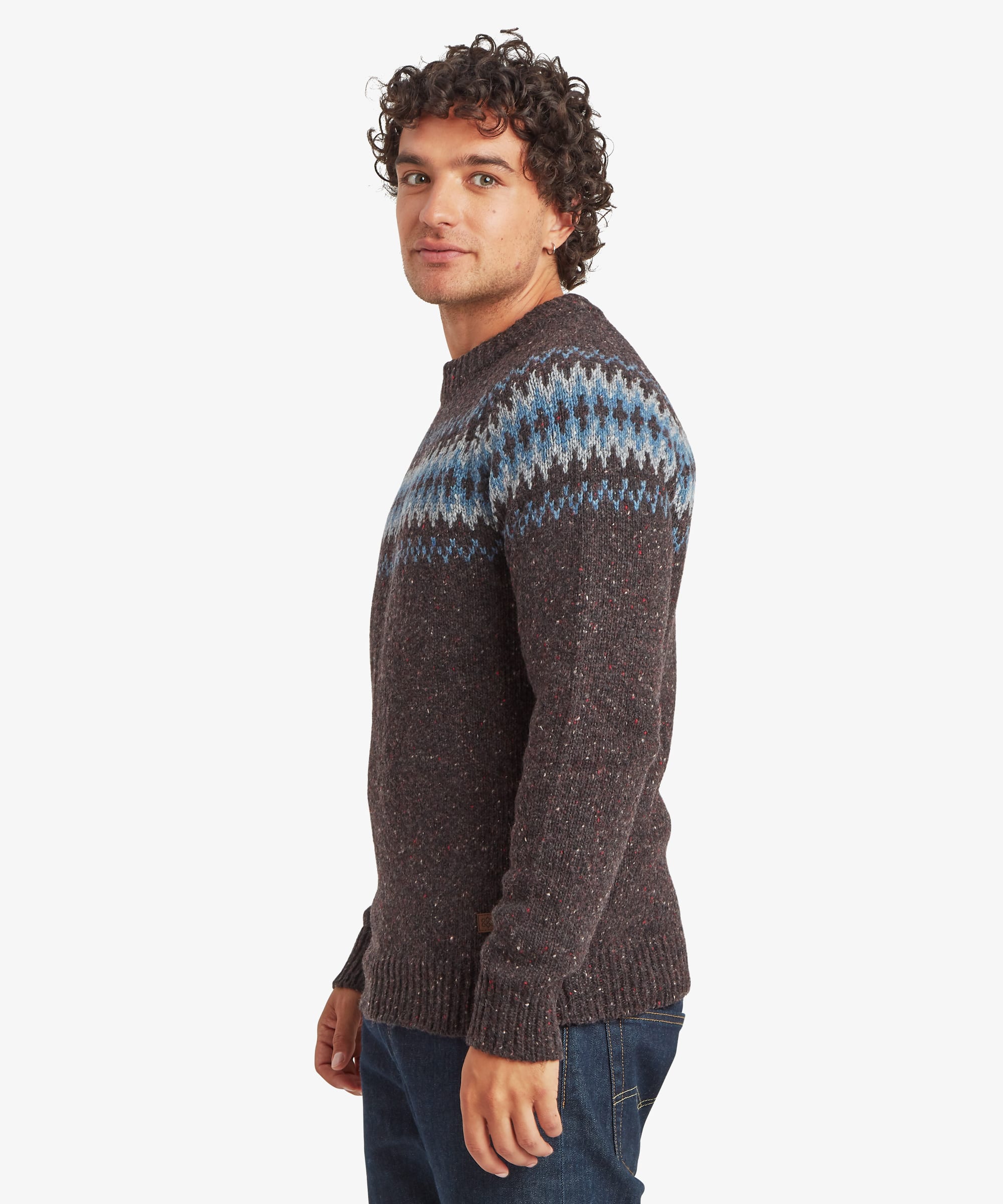 Side profile of a man wearing a charcoal black knit sweater with a Fair Isle-inspired patterned yoke in shades of blue, grey and white. The regular-fit sweater falls comfortably through the body and sleeves and is paired with blue jeans.