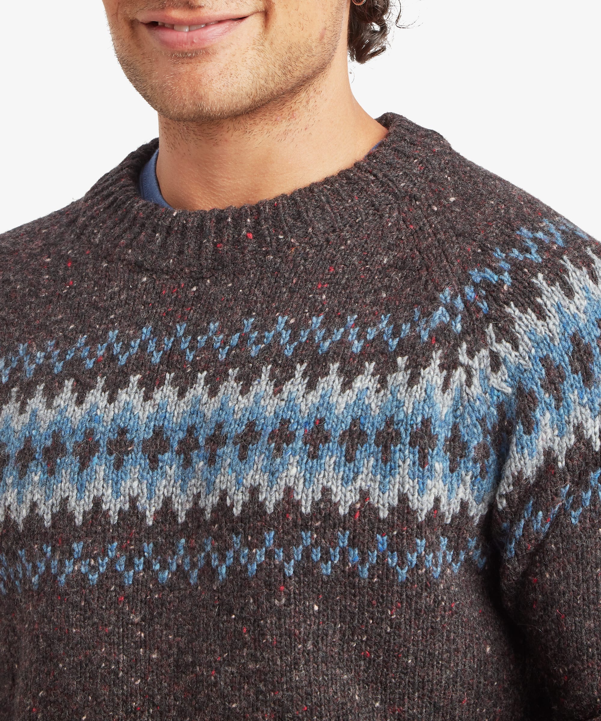 Close-up detail of a charcoal black crew neck sweater highlighting the textured knit construction and geometric Fair Isle-inspired pattern in blue, grey and white across the upper chest. Ribbed neckline detail is clearly visible.