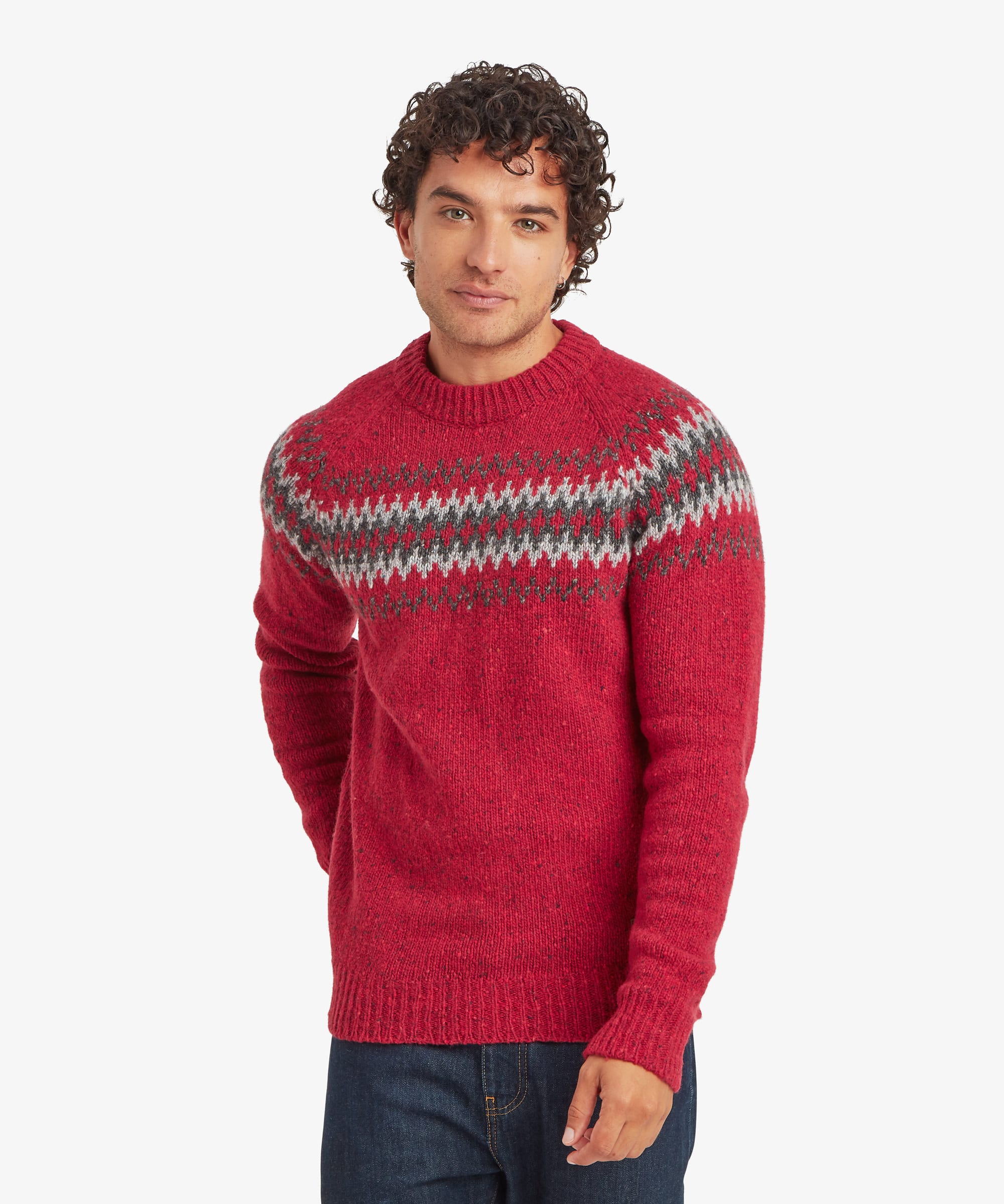 Man wearing a vibrant red crew neck knit sweater with a multi-coloured Fair Isle-inspired geometric pattern in grey, black and white across the chest and shoulders. He stands with a relaxed expression and hands behind his back. The regular-fit sweater is paired with blue jeans.