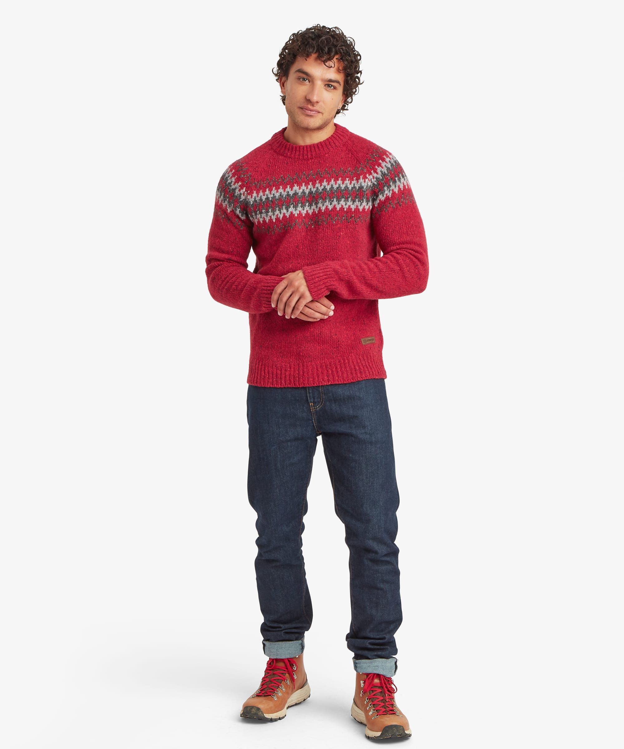 Full-length front view of a man wearing a red crew neck sweater featuring a geometric patterned yoke in grey, black and white tones. Styled with blue jeans and casual shoes, the sweater has a regular fit with ribbed neckline, cuffs and hem.