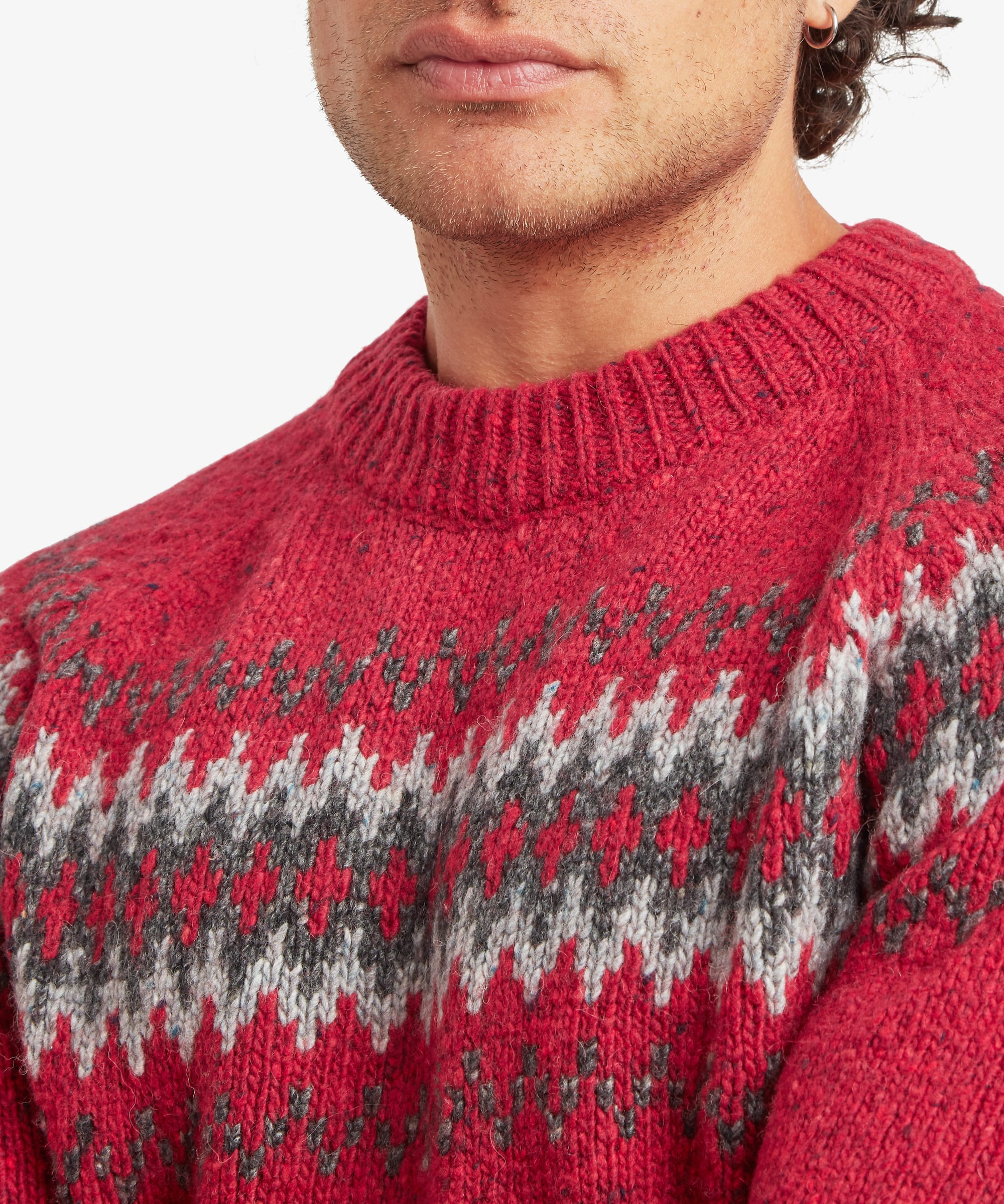 Close-up of a red crew neck sweater showcasing the intricate geometric Fair Isle-inspired pattern in grey, black and white. The ribbed neckline and textured knit fabric are shown in detail.