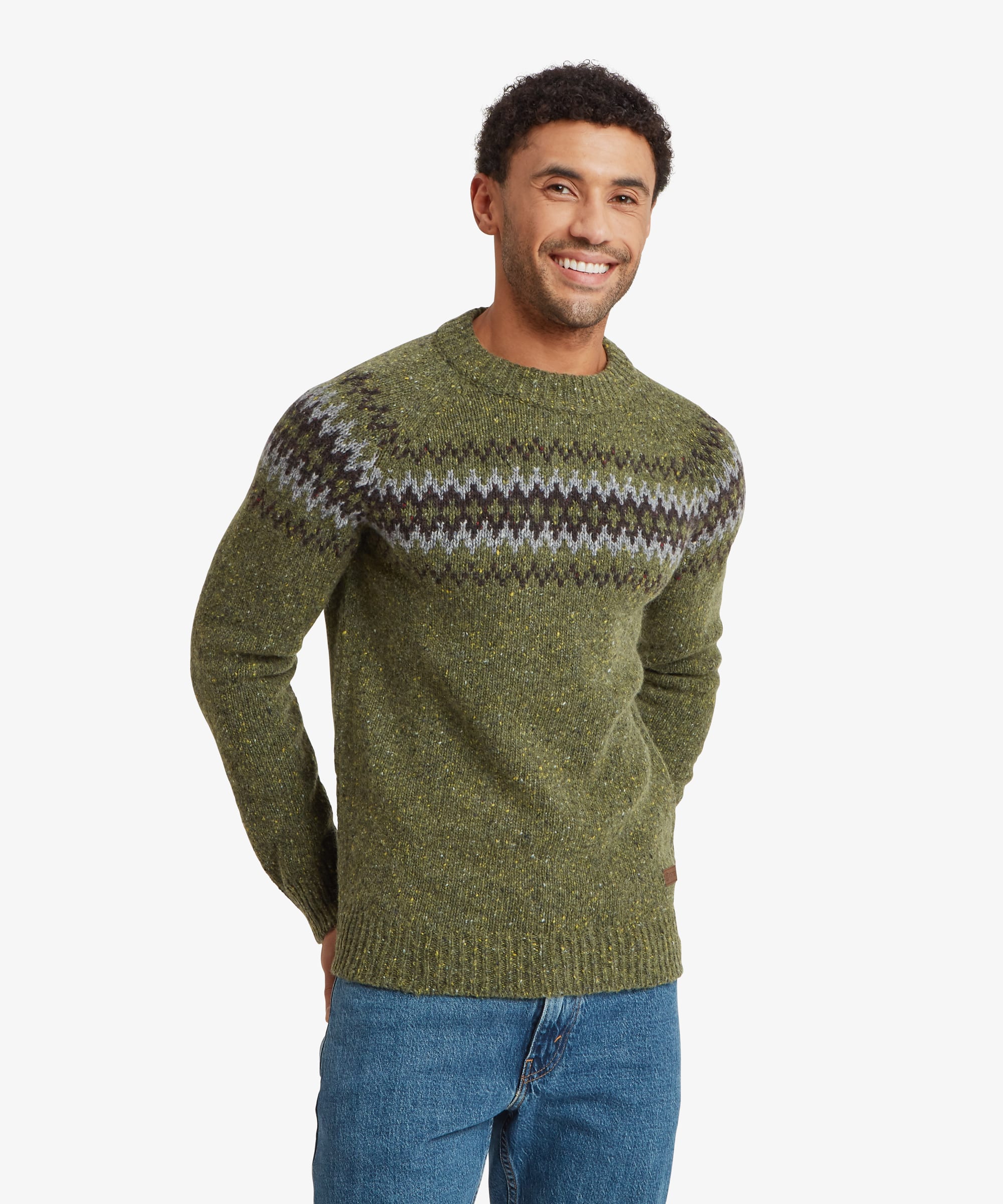 Smiling man wearing an olive green crew neck knit sweater with a contrasting Fair Isle-inspired geometric pattern in black, grey and cream across the chest and shoulders. The regular-fit sweater is paired with blue jeans and features ribbed cuffs, hem and neckline.