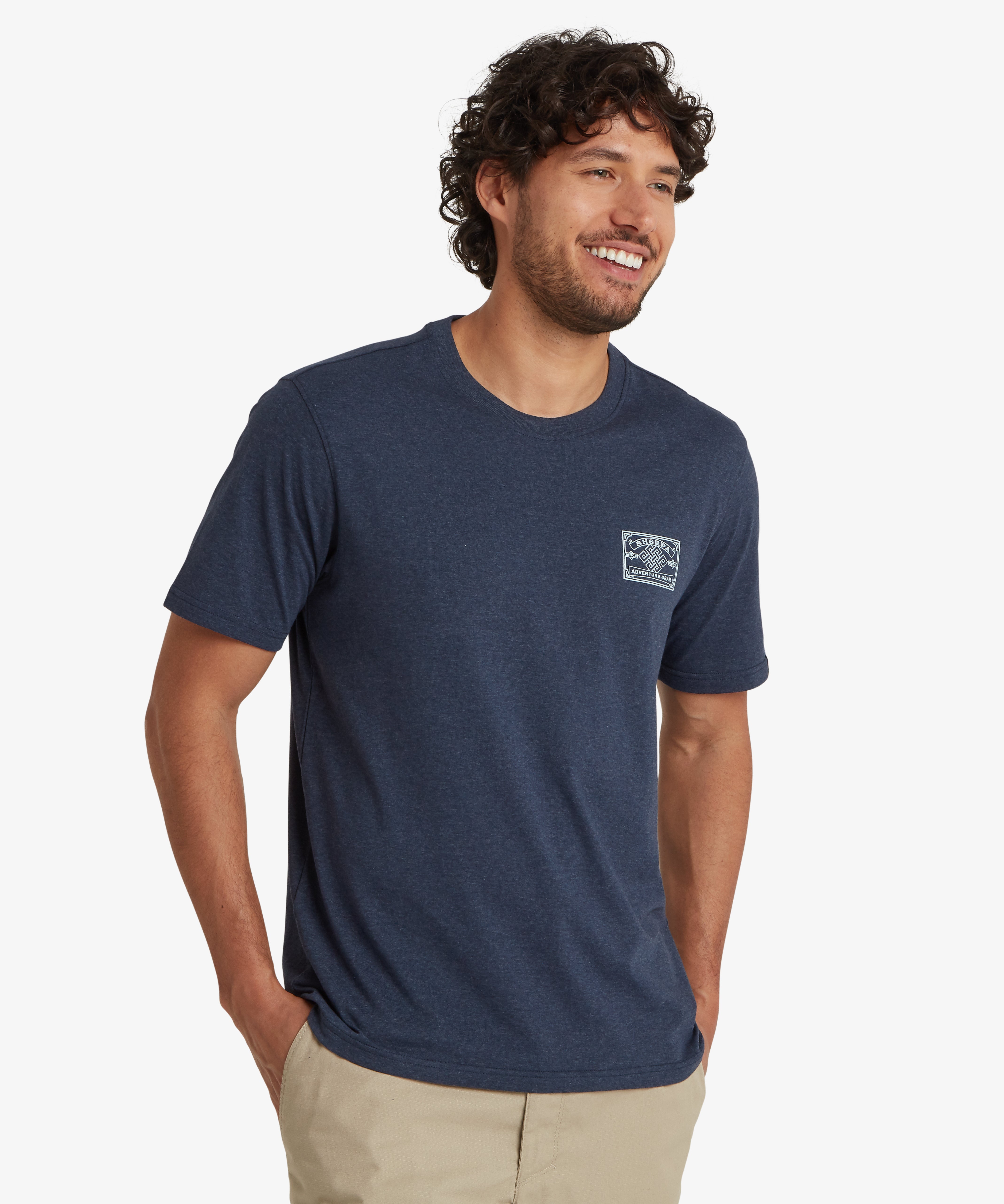 A mid shot of a man wearing a blue t-shirt with a white mountaineering inspired heritage patch printed on the left chest. At the centre of the patch is a Nepalese endless knot surrounded by the word "Sherpa Adventure Gear". The fabric of the T-shirts looks soft and has a flattering drape. The t-shirt is styled with a pair of beige shorts. The man is standing against a white background with both hand in his shorts pockets. The outfit is an ideal summer outfit for gentle outdoor pursuits.