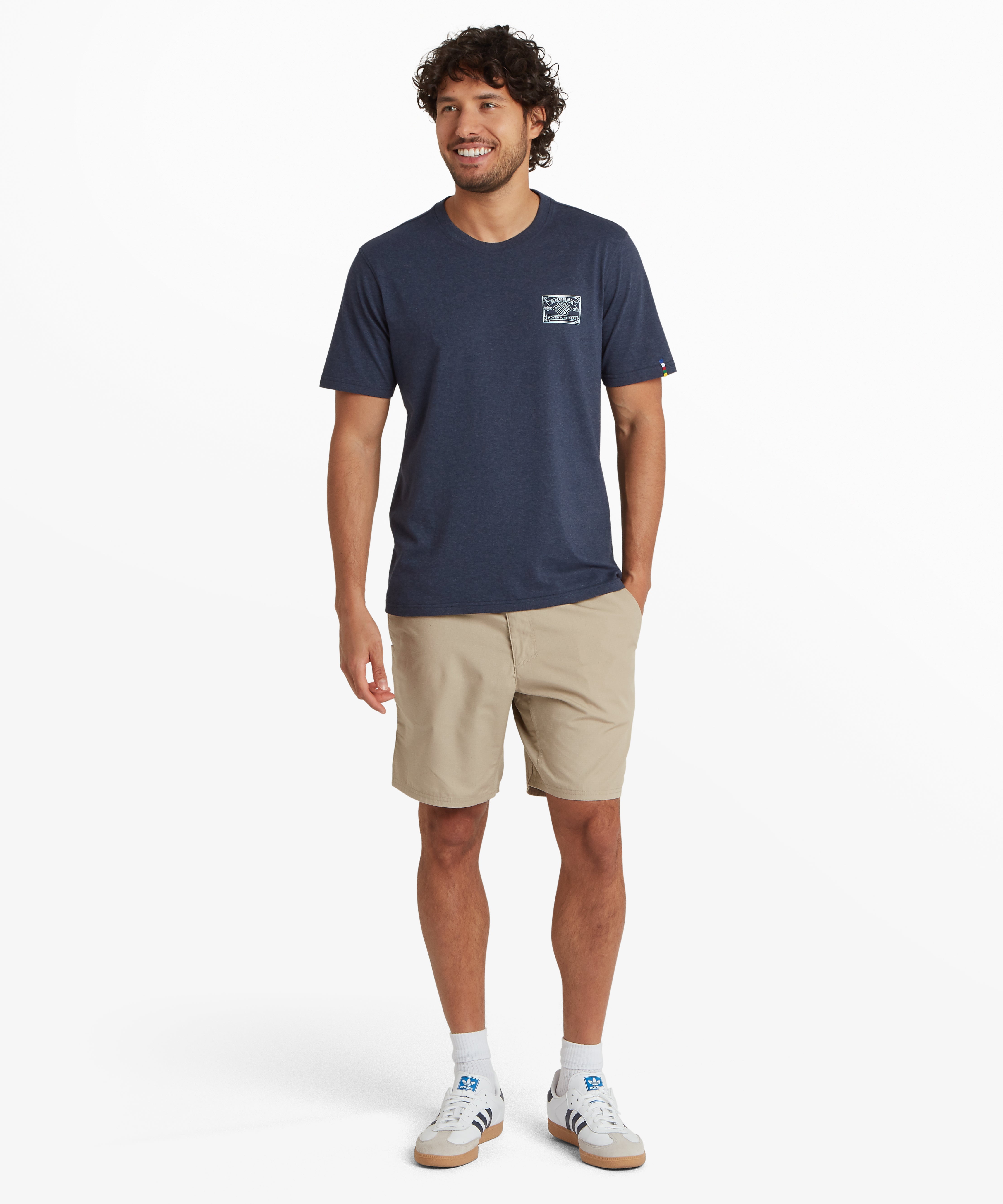 Ganzkšrperaufnahme eines Mannes, der ein blaues T-Shirt mit einem wei§en, vom Bergsteigen inspirierten Heritage-AufnŠher auf der linken Brust trŠgt. In der Mitte des AufnŠhers befindet sich ein nepalesischer Endlosknoten, umgeben vom Schriftzug ãSherpa Adventure GearÒ. Das T-Shirt wird mit einer beigen Shorts, wei§en Socken und wei§en Turnschuhen kombiniert. Der Mann steht vor einem wei§en Hintergrund und hat seine linke Hand in der Hosentasche. Das Outfit ist ideal fŸr leichte Outdoor-AktivitŠten.