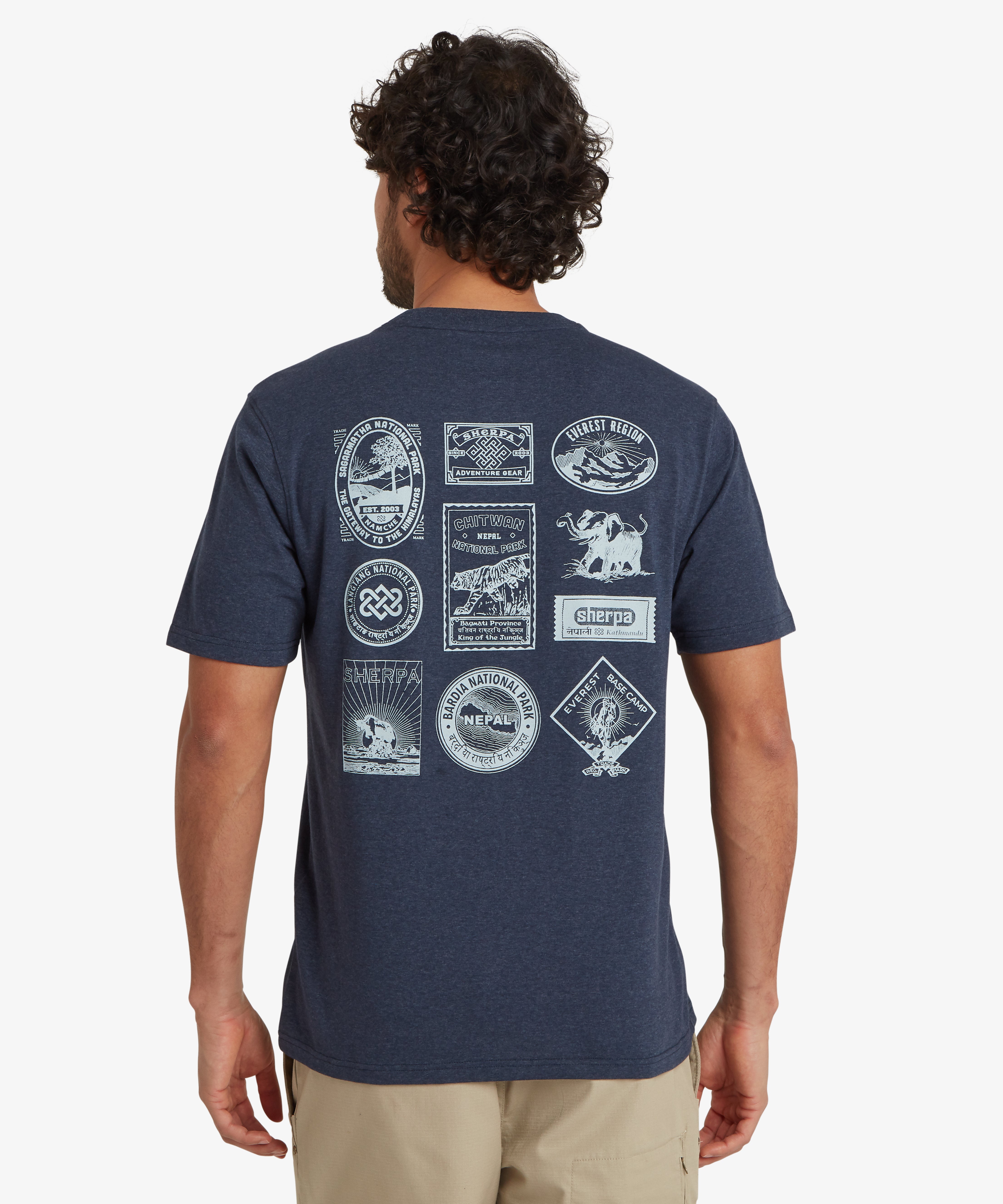 A back view of a man wearing a blue short sleeve t-shirt. The t-shirt has 10 Nepal inspired travel and heritage patches printed onto the back in white. Many of them depict mountain ranges or national parks in Nepal as well as showing the Sherpa Adventure Gear logo. The t-shirt is styled with a pair of beige shorts with a zipped pocket on the side creating a casual summer outfit for outdoor walking or activities. The man is standing against a white background with his hands down by his sides.