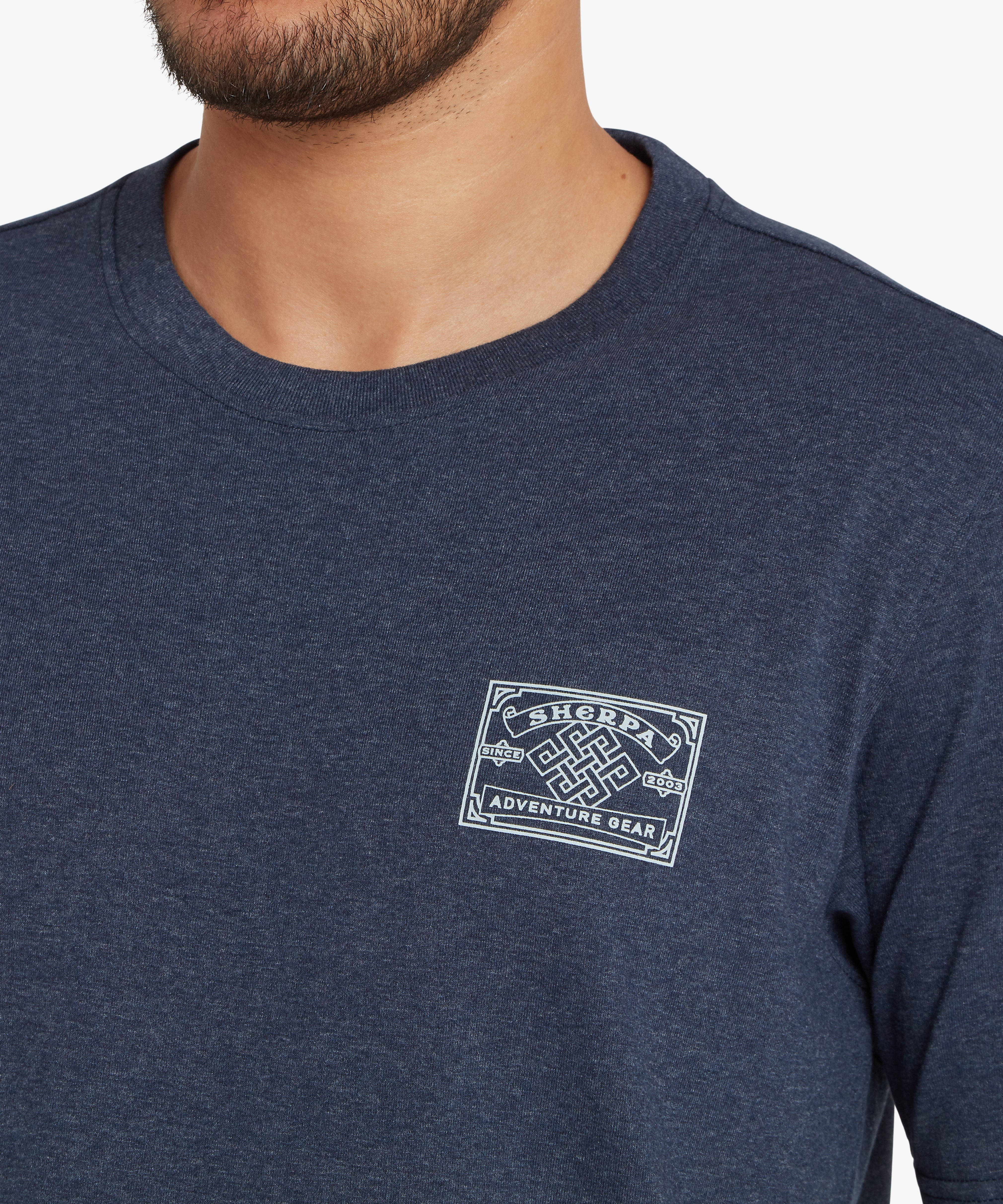 Nahaufnahme eines Mannes, der ein blaues T-Shirt mit einem wei§en AufnŠher im Heritage-Stil auf der linken Brust trŠgt, der vom nepalesischen Bergsteigen inspiriert ist. Der AufnŠher zeigt in der Mitte einen Endlosknoten, Ÿber und unter dem die Worte ãSherpa Adventure GearÓ aufgedruckt sind.  Zu beiden Seiten des Knotens steht der Schriftzug ãsince 2003Ó. Der Mann steht vor einem wei§en Hintergrund.
