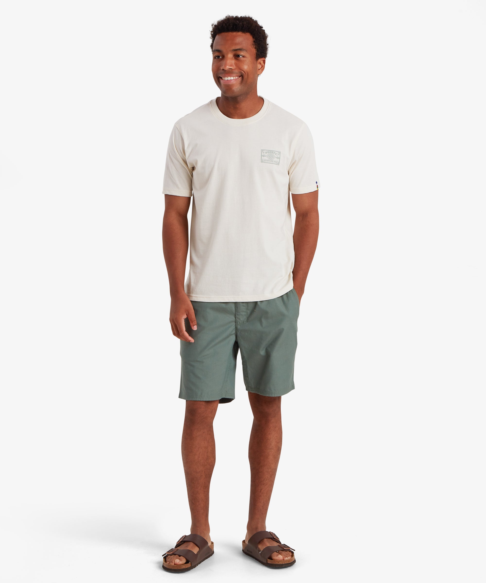 Ein Mann trŠgt ein wei§es T-Shirt mit einem auf der linken Brust aufgedruckten Heritage-AufnŠher, der vom Bergsteigen in Nepal inspiriert ist. Das T-Shirt wird mit einer hellgrŸnen Shorts, die Ÿber dem Knie endet, und braunen Sandalen kombiniert. Das T-Shirt hat einen kleinen AufnŠher auf dem linken €rmel in den Farben nepalesischer Gebetsfahnen. Der Mann steht vor einem wei§en Hintergrund, lŠchelt und hat eine Hand in der Tasche.