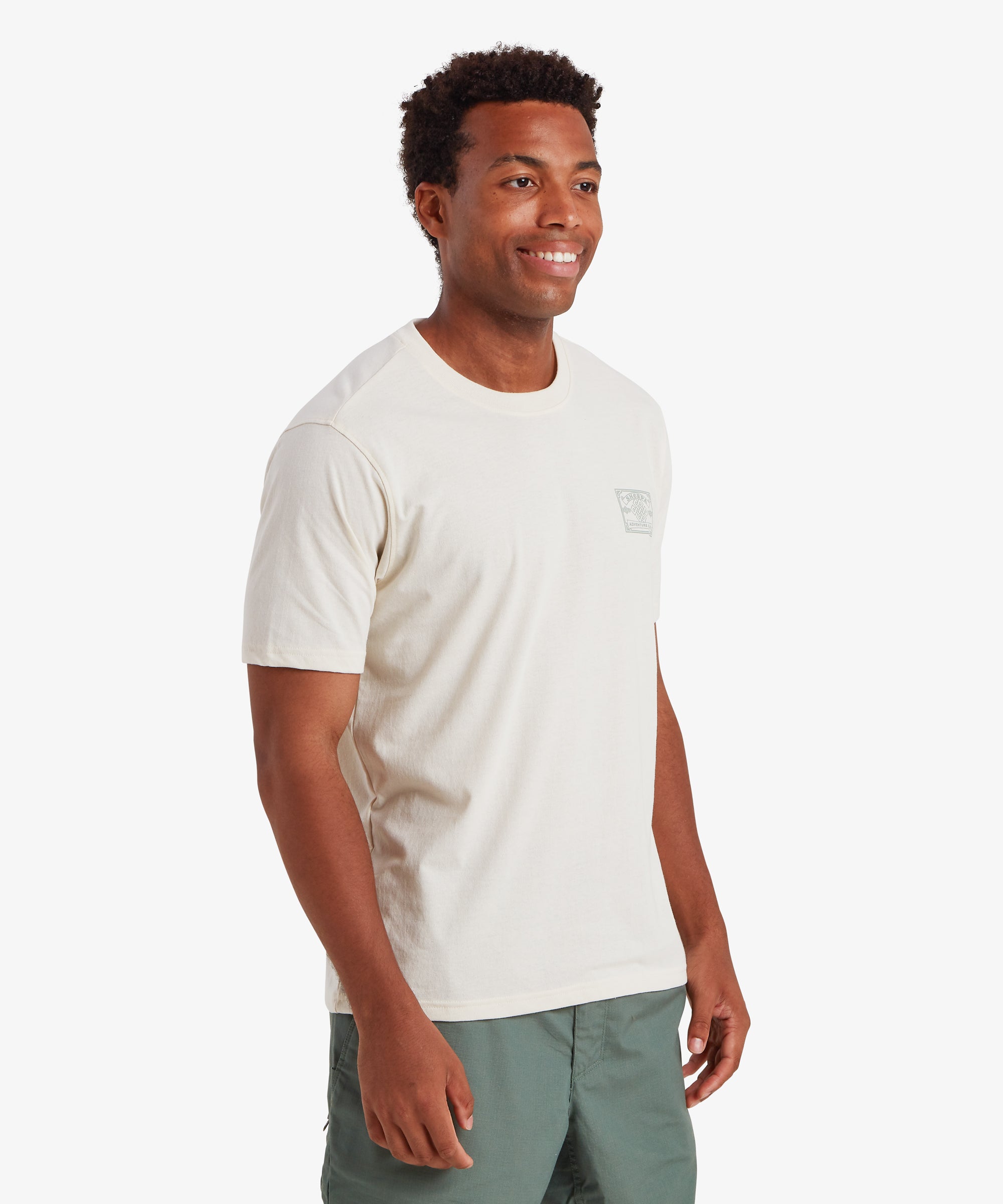 Ein Mann trŠgt ein wei§es Kurzarm-T-Shirt mit einem hellgrŸnen AufnŠher im Heritage-Stil auf der linken Brust, der vom Bergsteigen inspiriert ist. Das T-Shirt wird mit hellgrŸnen Shorts kombiniert. Das Outfit ist ideal fŸr alltŠgliche AktivitŠten bei warmem Wetter. Der Mann ist nach links gedreht und steht mit beiden HŠnden an den Seiten.