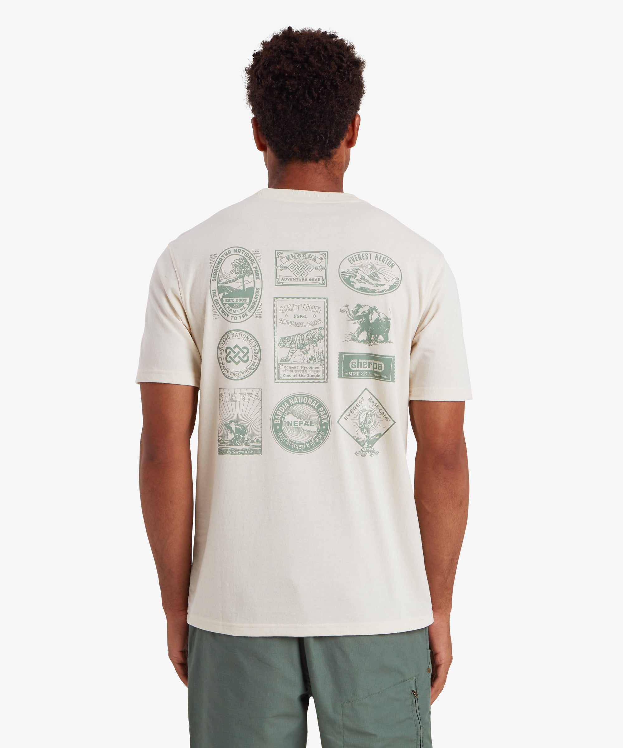 RŸckansicht eines Mannes, der ein wei§es Kurzarm-T-Shirt trŠgt. Auf dem RŸcken des T-Shirts sind 10 von Nepal inspirierte Reise- und Heritage-AufnŠher in hellgrŸner Farbe aufgedruckt. Viele davon zeigen GebirgszŸge oder Nationalparks in Nepal sowie das Logo von Sherpa Adventure Gear. Das T-Shirt wird mit hellgrŸnen Shorts mit einer Rei§verschlusstasche auf der RŸckseite kombiniert. Der Mann steht vor einem wei§en Hintergrund, die HŠnde an den Seiten.