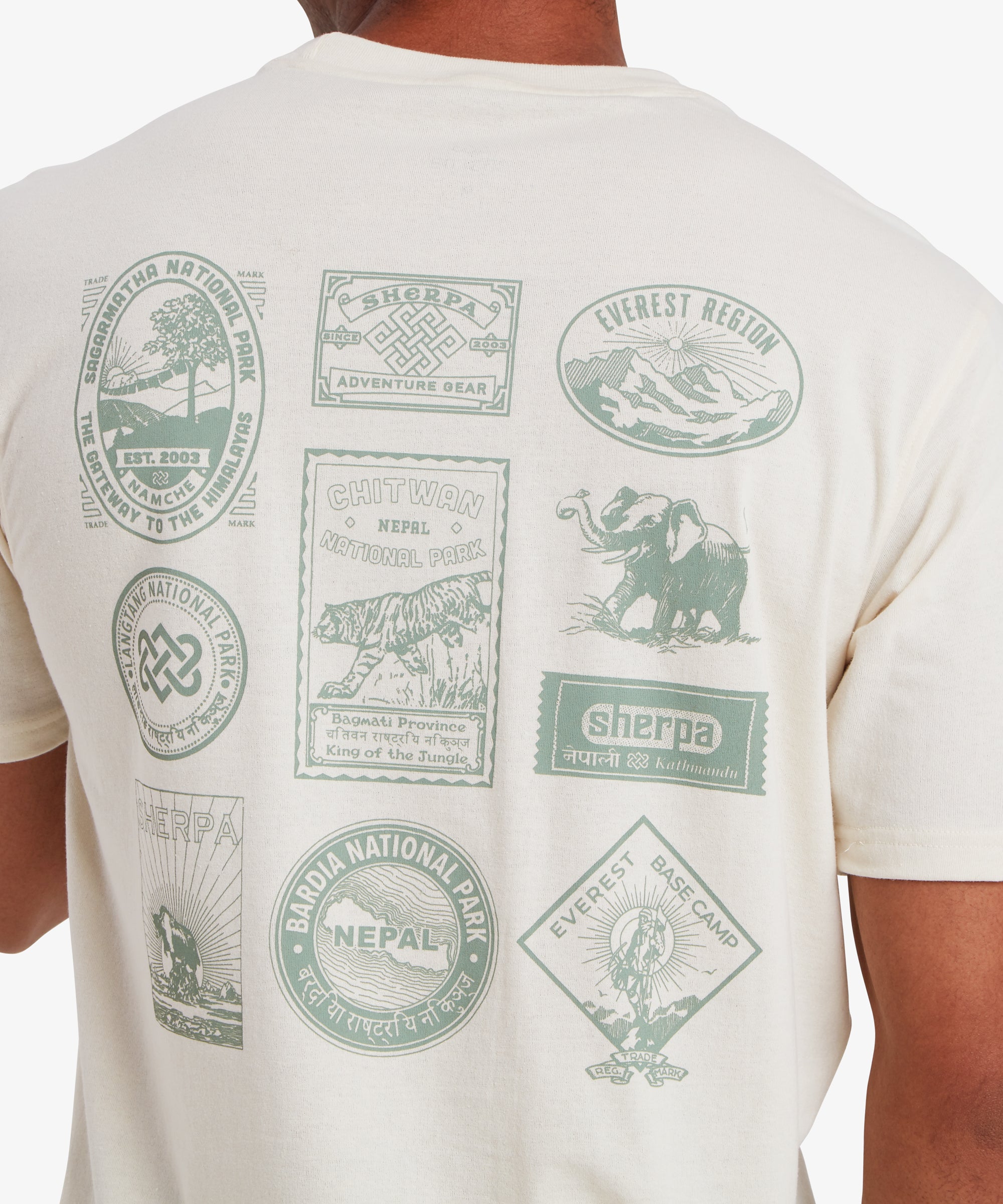 Nahaufnahme des RŸckens eines Mannes, der ein wei§es Kurzarm-T-Shirt trŠgt. Auf dem T-Shirt sind 10 von Nepal inspirierte Reise- und Heritage-AufnŠher in hellgrŸner Farbe aufgedruckt. Viele davon zeigen GebirgszŸge oder Nationalparks in Nepal sowie das Logo von Sherpa Adventure Gear.