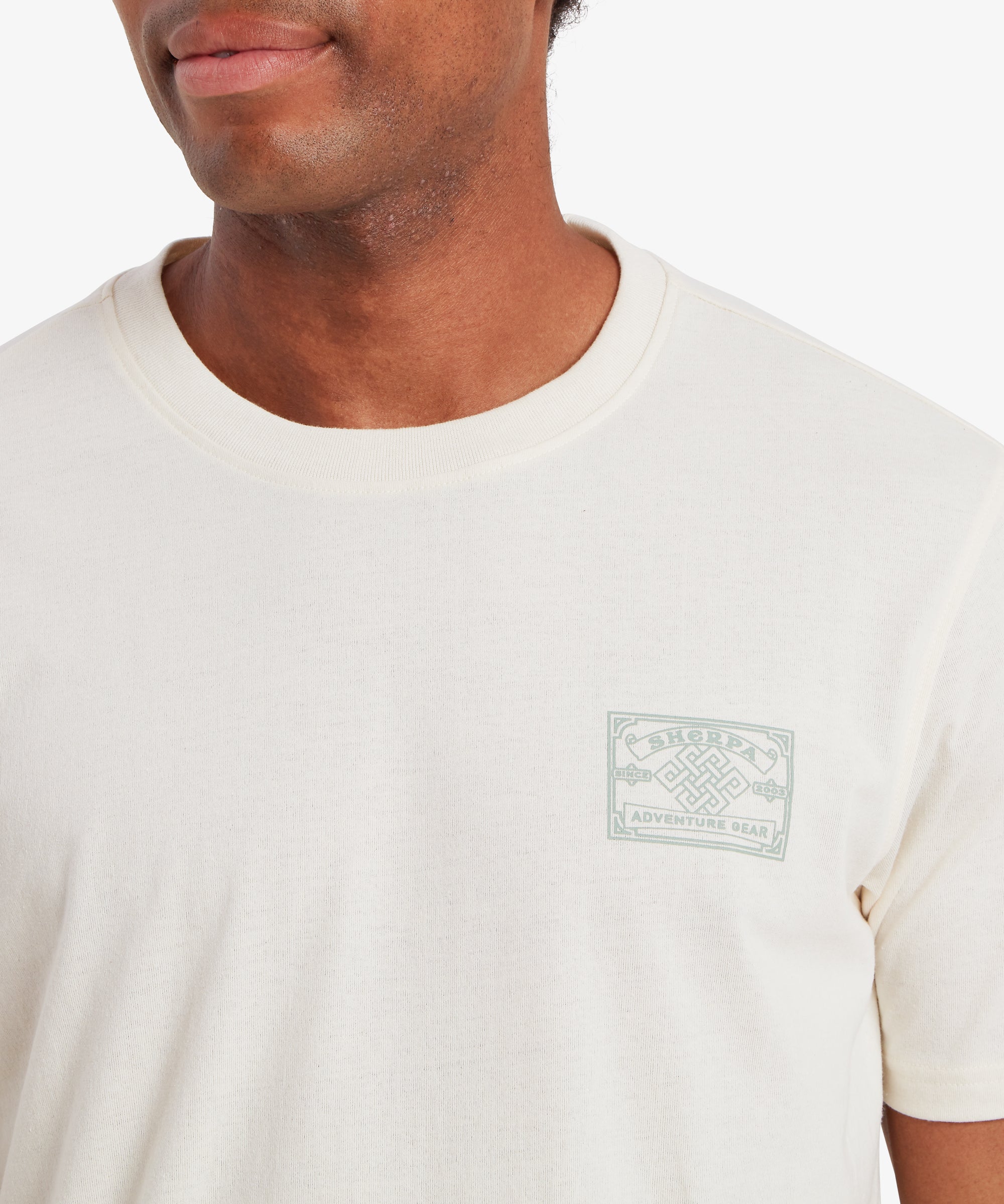 Nahaufnahme eines Mannes, der ein wei§es T-Shirt mit einem auf der linken Brustseite aufgedruckten AufnŠher mit einem Motiv aus der nepalesischen Bergsteigerkultur trŠgt. Der Mann steht lŠchelnd vor einem wei§en Hintergrund.