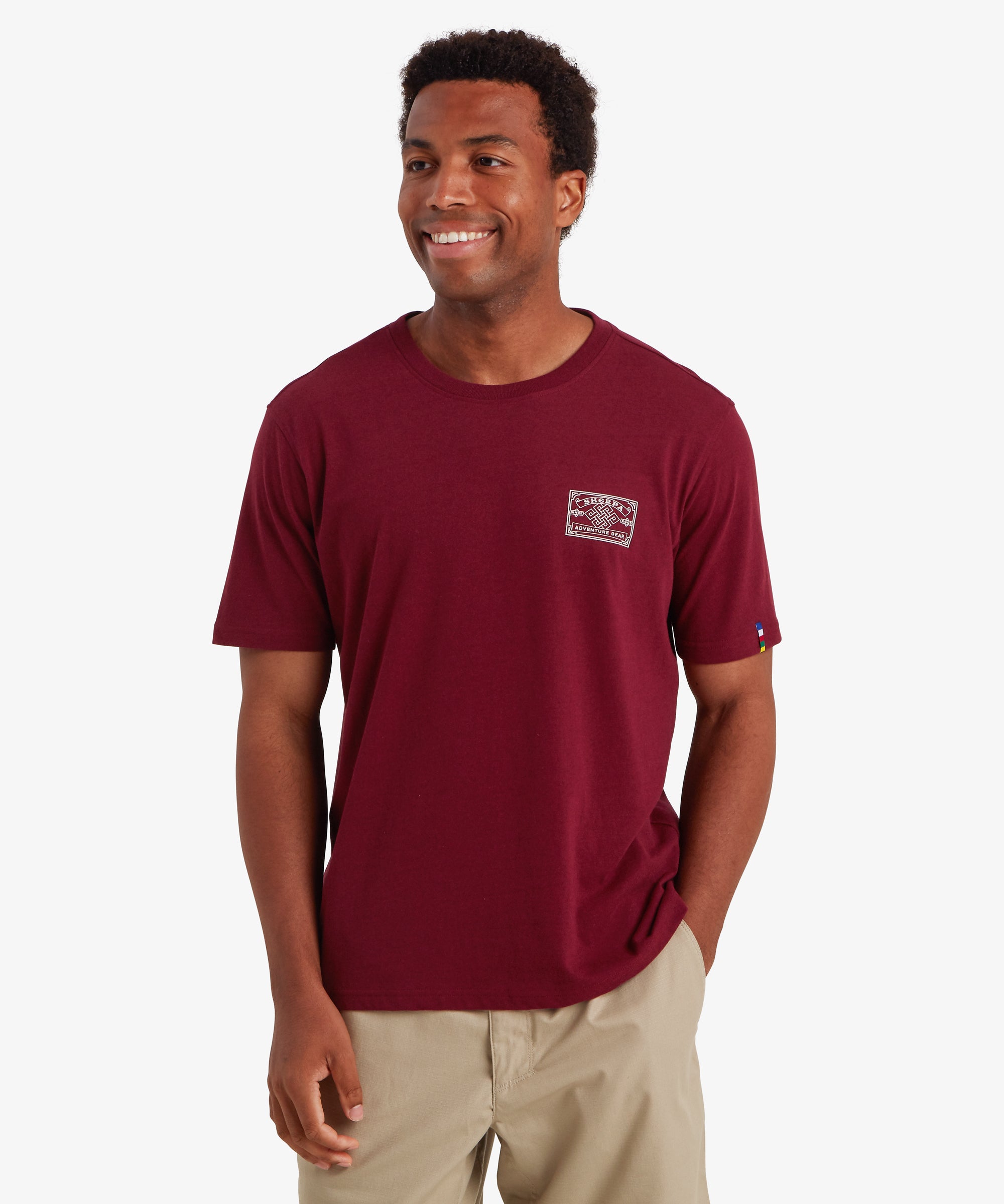 Ein Mann trŠgt ein rotes T-Shirt mit einem wei§en AufnŠher im Heritage-Stil auf der linken Brust, der vom Bergsteigen inspiriert ist. Das T-Shirt wird mit einer beigen Shorts kombiniert. Das T-Shirt hat einen kleinen AufnŠher auf dem linken €rmel in den Farben der nepalesischen Gebetsfahnen: grŸn, blau, wei§, rot und gelb. Der Mann steht vor einem wei§en Hintergrund und hat seine linke Hand in der Hosentasche. Das Outfit ist ideal fŸr den Sommer.