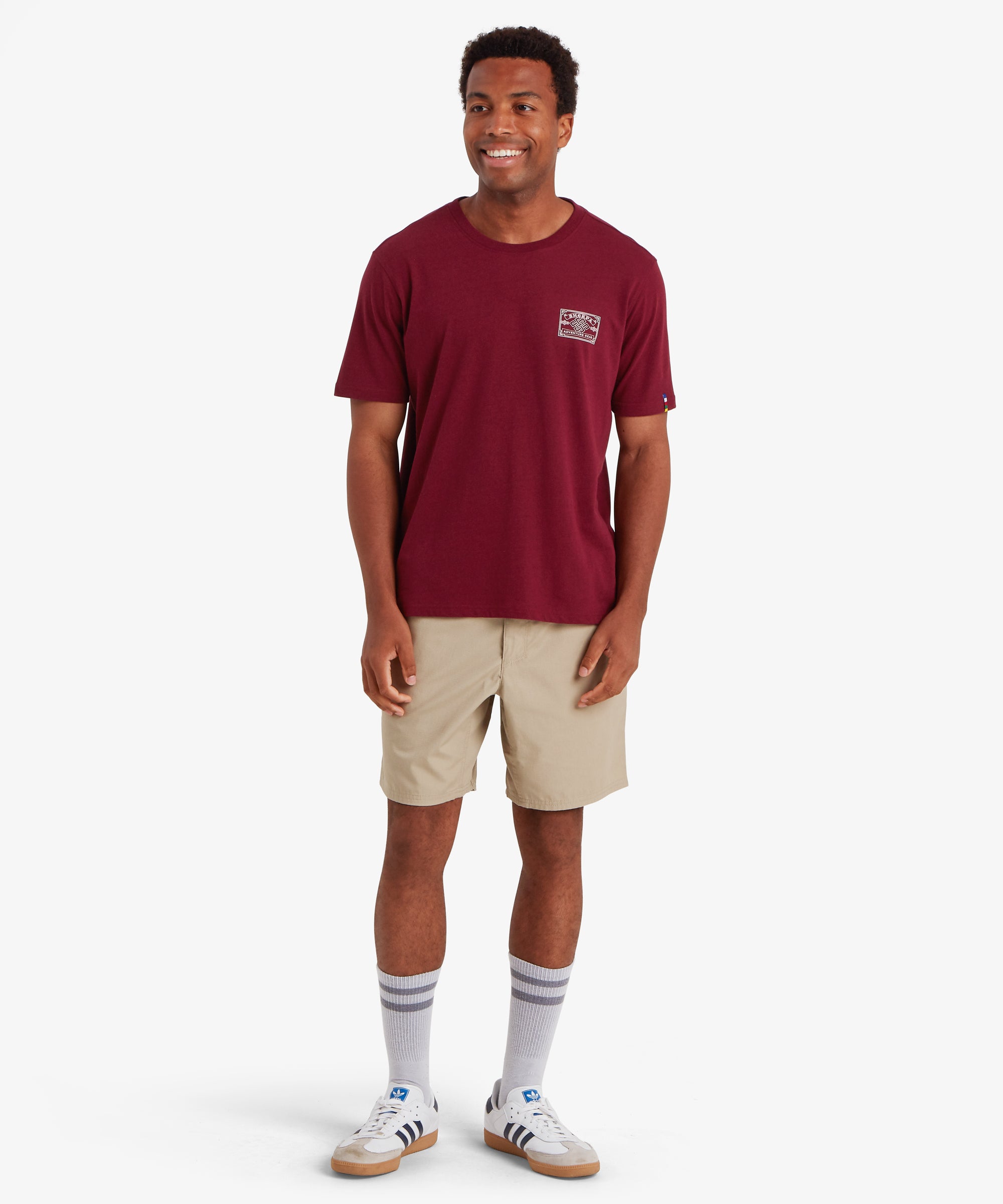 Man wearing a red t-shirt with a Nepal mountaineering inspired heritage patch printed on the left chest. The classic fit t-shirt is styled with a pair of above the knee beige shorts, white socks and white trainers. The t-shirt has a small pip on the left sleeve in the colors of Nepalese prayer flags. The man is standing against a white background smiling with both hands down by his side. The outfit is perfect for casual, summer wear.