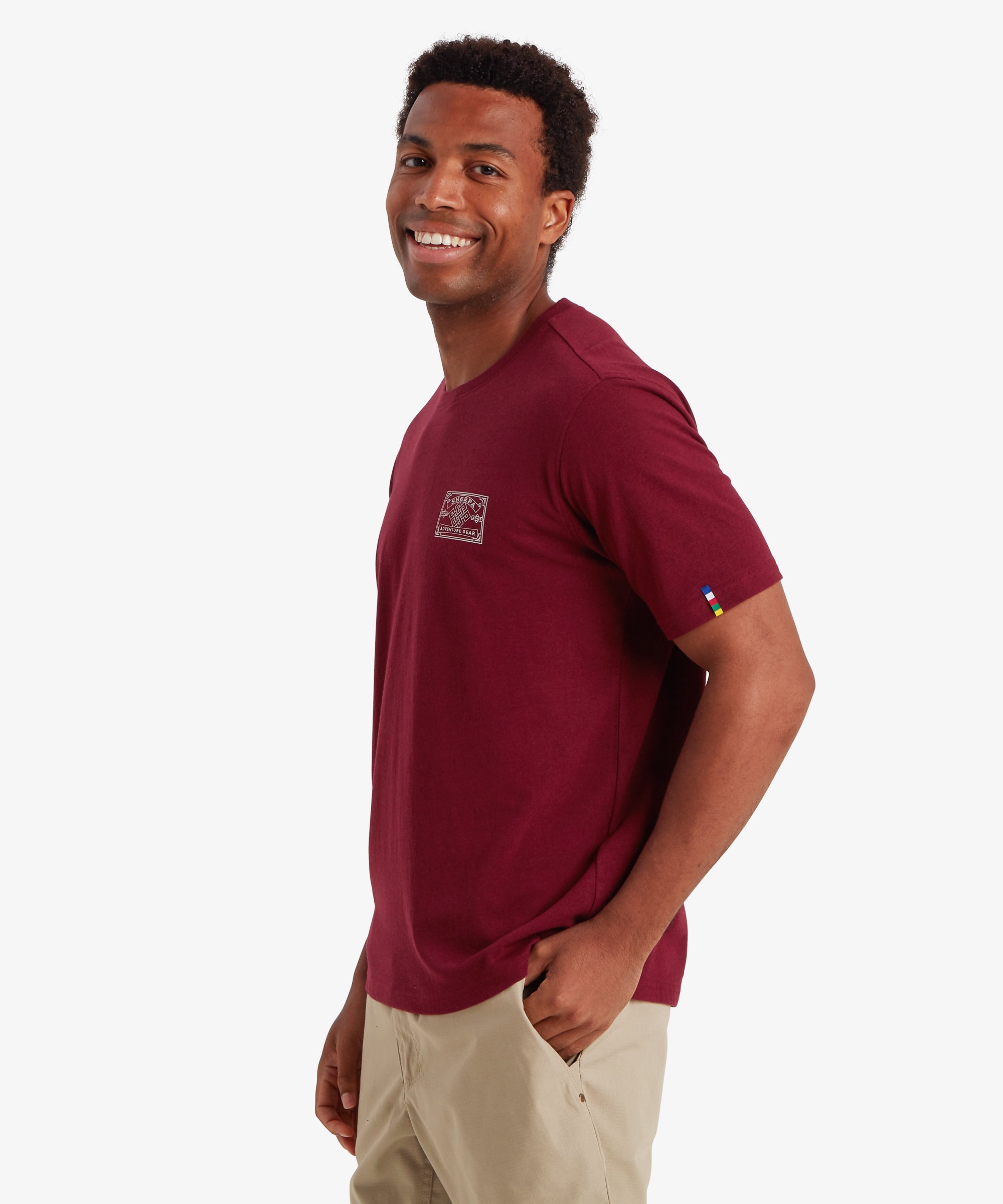 Mann mit rotem T-Shirt mit einem wei§en AufnŠher im Heritage-Stil auf der linken Brust, der vom Bergsteigen inspiriert ist. Das T-Shirt wird mit einer beigen Shorts kombiniert. Das T-Shirt hat einen kleinen AufnŠher auf dem linken €rmel in den Farben nepalesischer Gebetsfahnen. Der Mann steht vor einem wei§en Hintergrund und hat seinen Kšrper nach rechts gedreht. Er schaut leicht Ÿber seine Schulter und lŠchelt. Das Outfit ist ideal fŸr den Sommer und den Freizeitbereich.