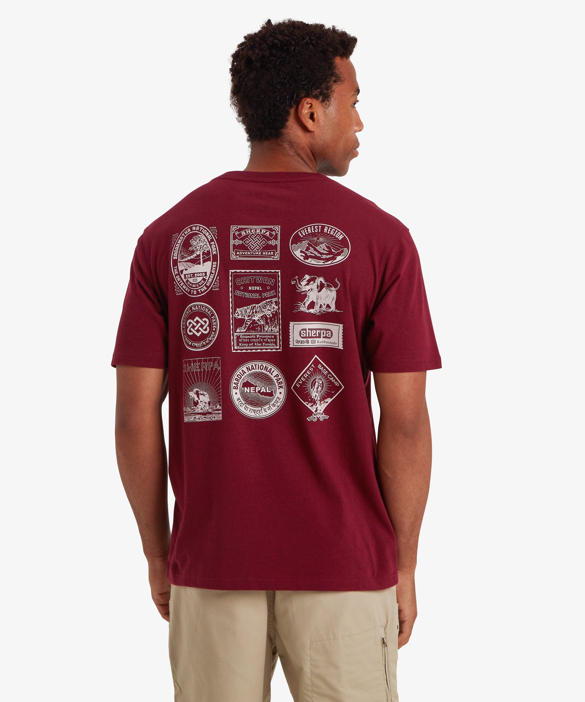RŸckansicht eines Mannes, der ein rotes Kurzarm-T-Shirt trŠgt. Auf dem RŸcken des T-Shirts sind 10 von Nepal inspirierte Reise- und Heritage-AufnŠher in Wei§ aufgedruckt. Viele davon zeigen GebirgszŸge oder Nationalparks in Nepal sowie das Logo von Sherpa Adventure Gear. Das T-Shirt wird mit einer beigen Shorts mit Rei§verschlusstasche an der Seite kombiniert. Der Mann steht vor einem wei§en Hintergrund, die HŠnde an den Seiten.